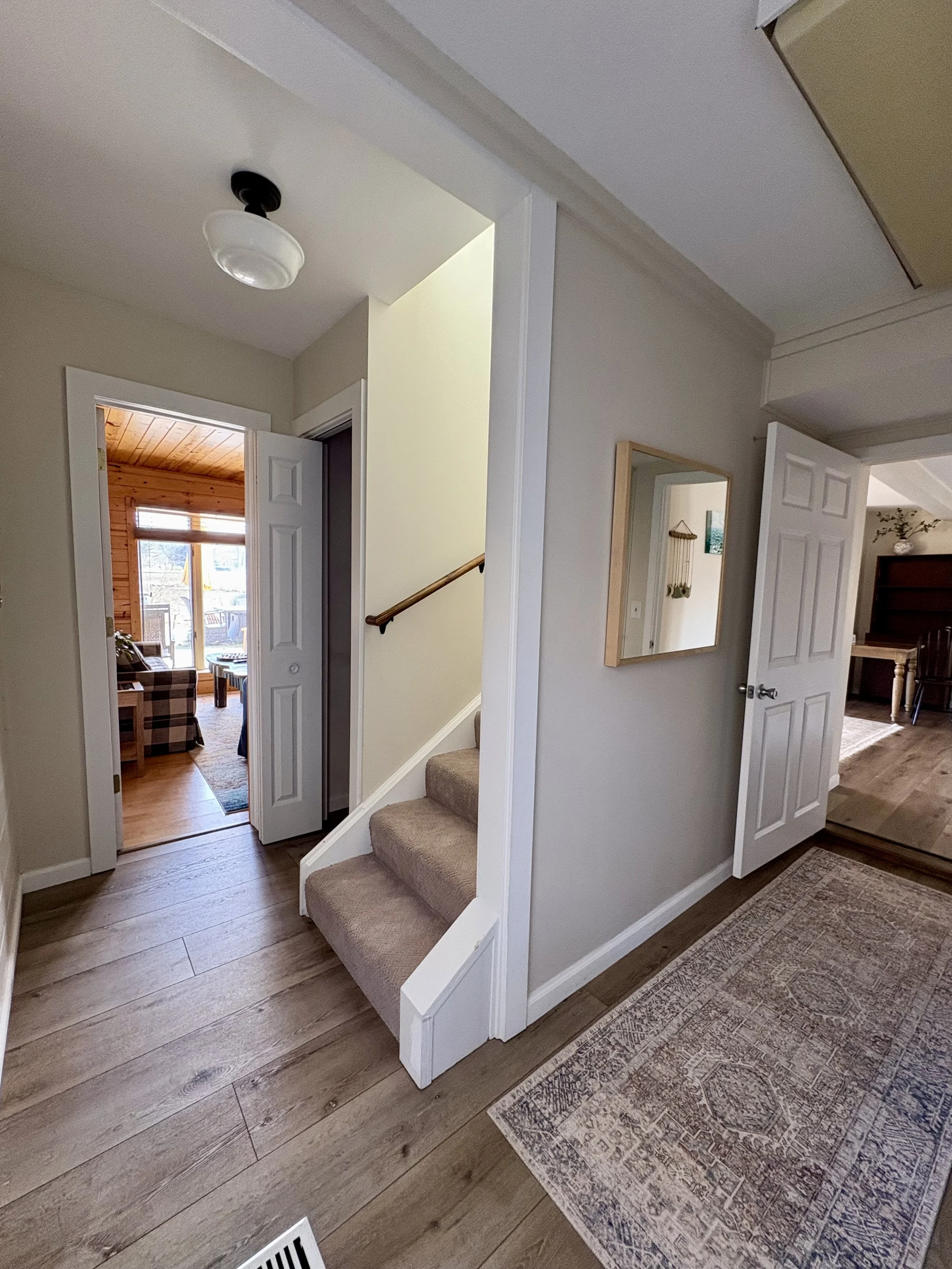 View of an interior home hallway with open doors leading to a living room with a wooden wall and large window, a staircase with carpeted steps, and a room with a dark wooden dresser and piano; hardwood floors, beige walls, and a ceiling with a light 