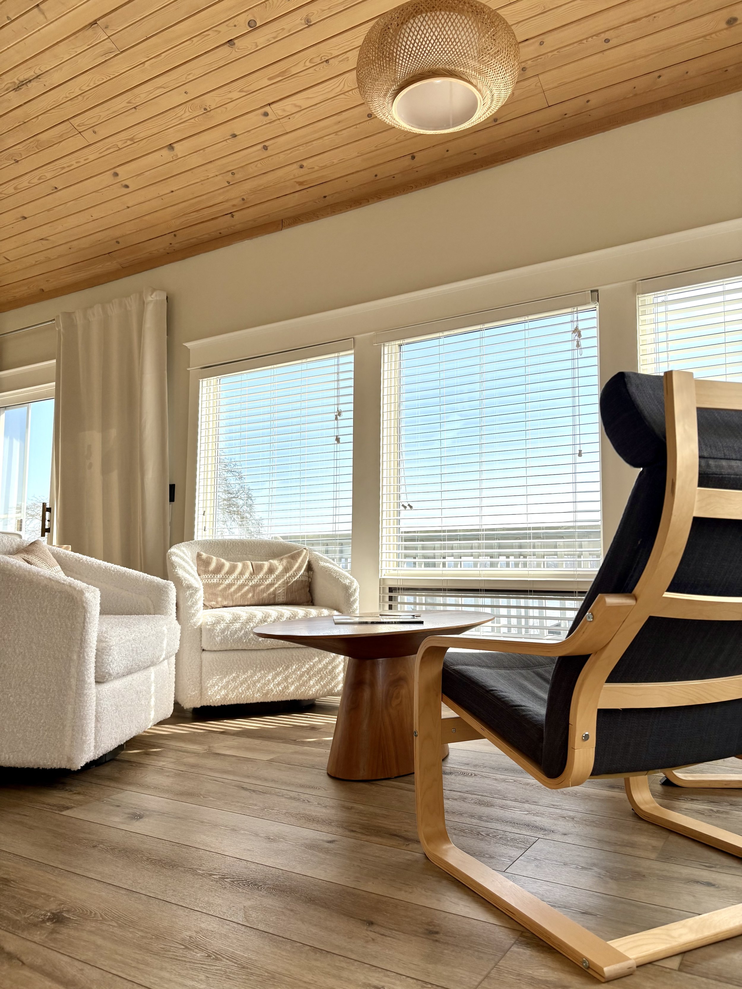 Living room with wooden ceiling, large windows with blinds, white curtained window to the left, white textured sofas, a wooden side table, and a black cushioned wooden armchair.