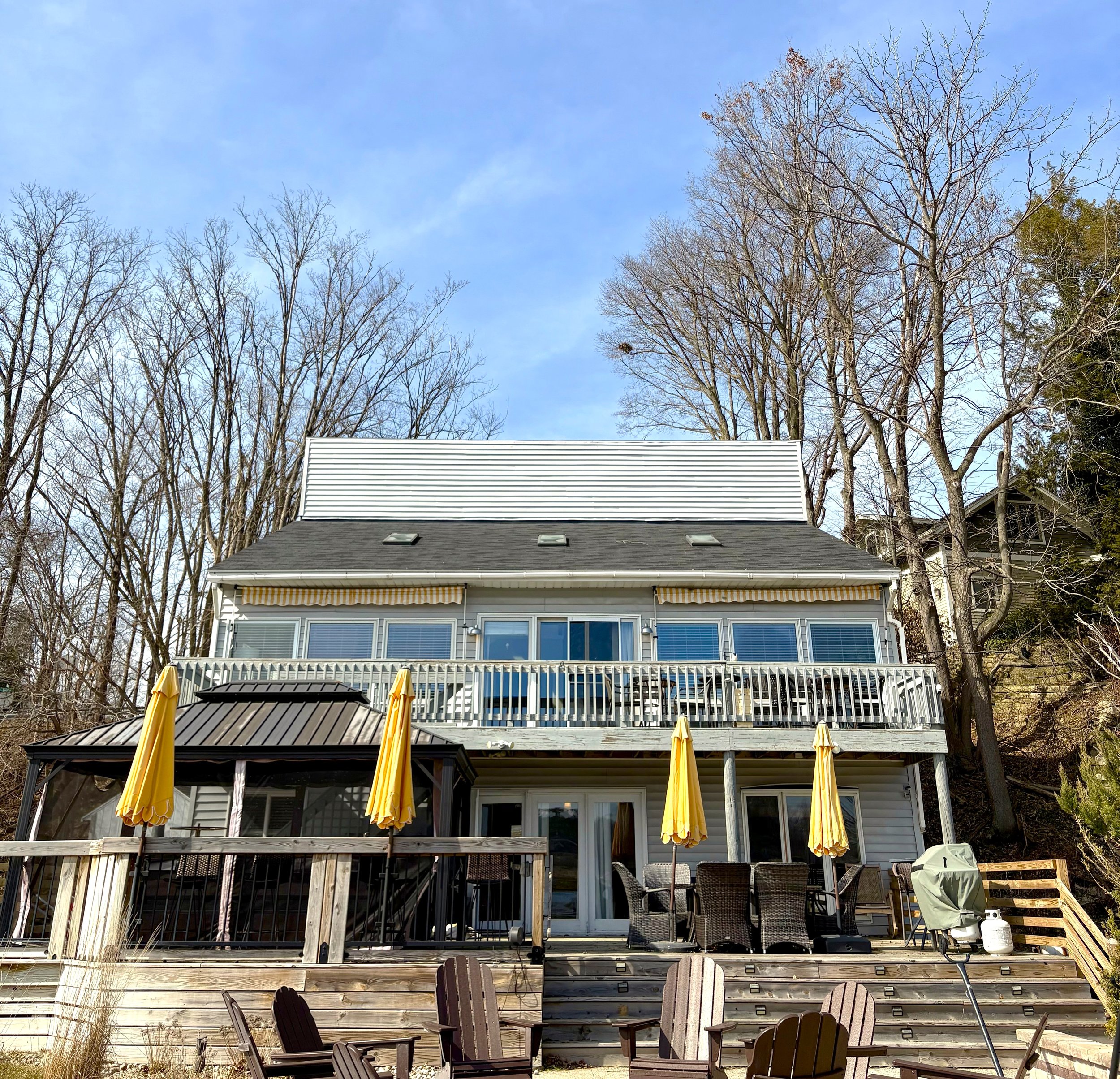 Backyard of a house featuring a wooden deck with patio furniture, yellow umbrellas, and a barbecue grill, with trees and a clear blue sky in the background.