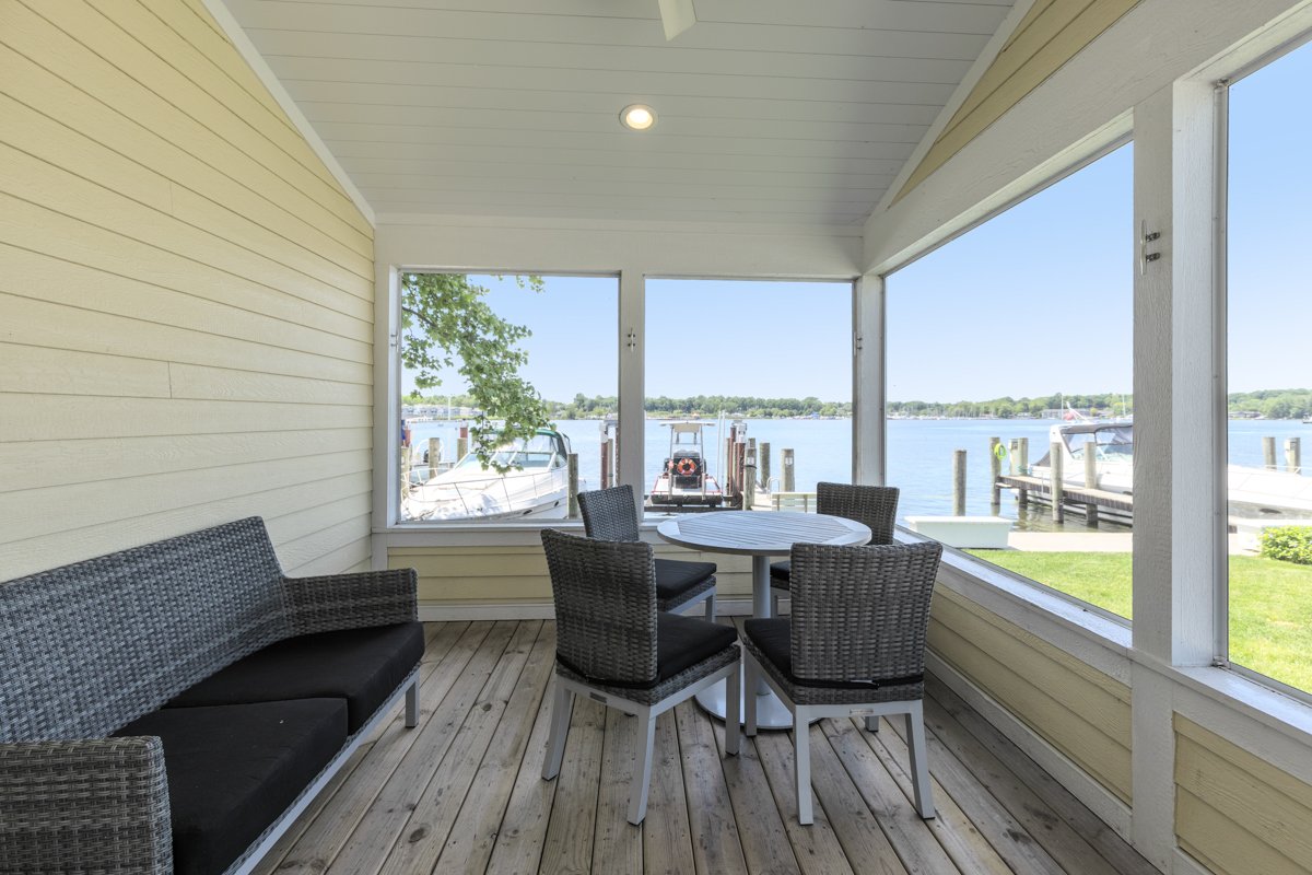 A screened porch overlooking a body of water with boats docked outside and seating including a wicker bench, a round table, and four chairs.