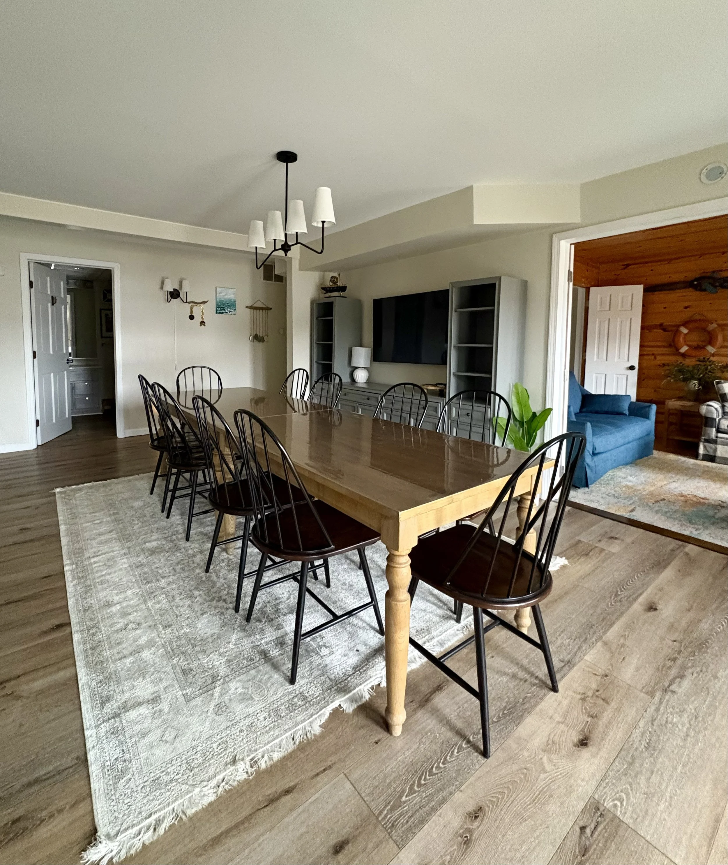 A spacious dining room with a large wooden table and black chairs, a patterned area rug, and a living area with a blue sofa and wooden paneling in the background.