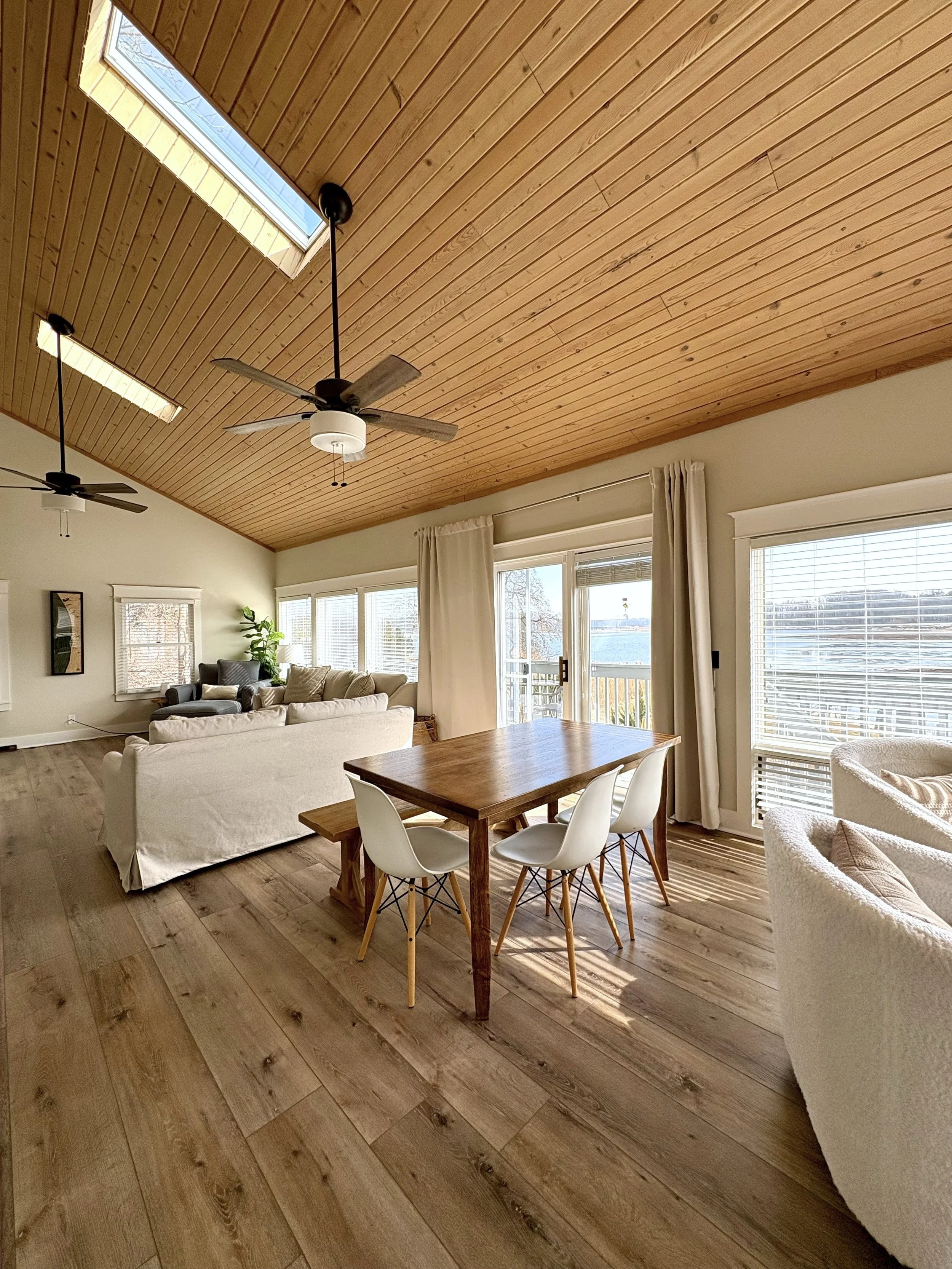Interior view of a living room and dining area with a wood-paneled vaulted ceiling, skylights, ceiling fans, large windows, and a sliding door leading outside. The space is furnished with a white sofa, a wooden dining table with white chairs, and a c