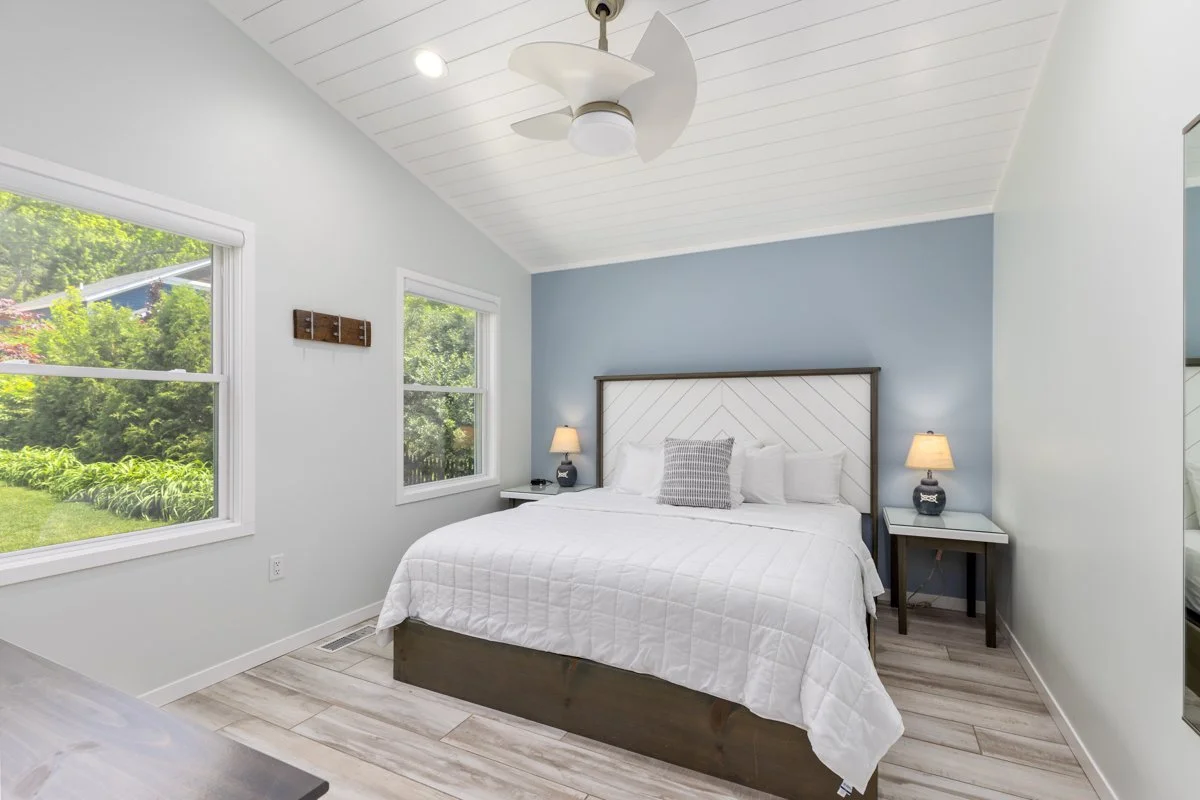 A bedroom with a white bed, two nightstands with lamps, and two windows showing a green outdoor landscape, light blue and white walls, wooden flooring, and a ceiling fan.