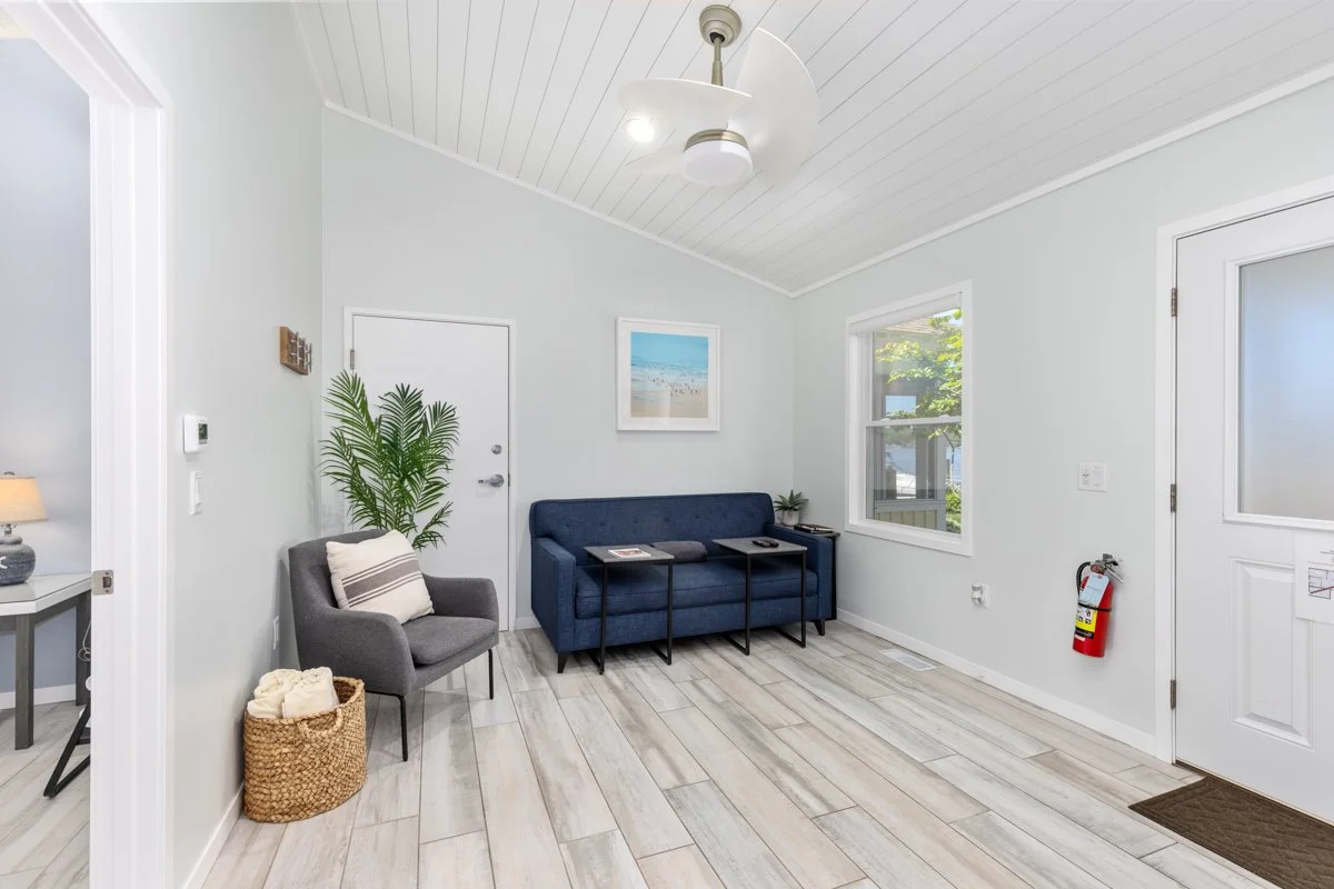 A bright living room with white walls and light-colored wood floor, furnished with a gray armchair, a blue sofa, and three black side tables. There is a large window letting in natural light, a ceiling fan, and a fire extinguisher next to the door.