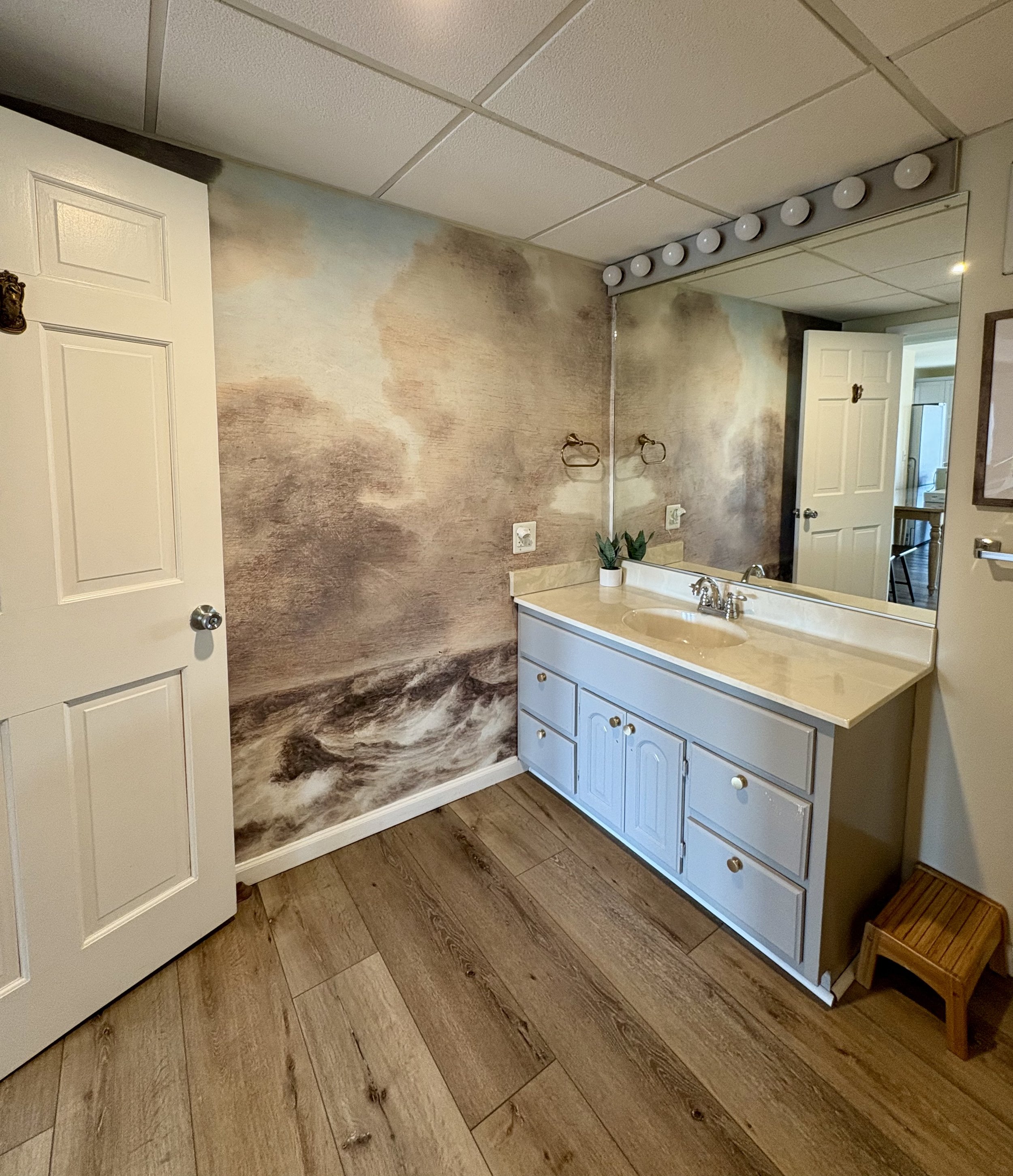 Bathroom with a large mirror, white vanity with a beige countertop, small potted plant, towel rings, and wall mural of a stormy ocean scene.