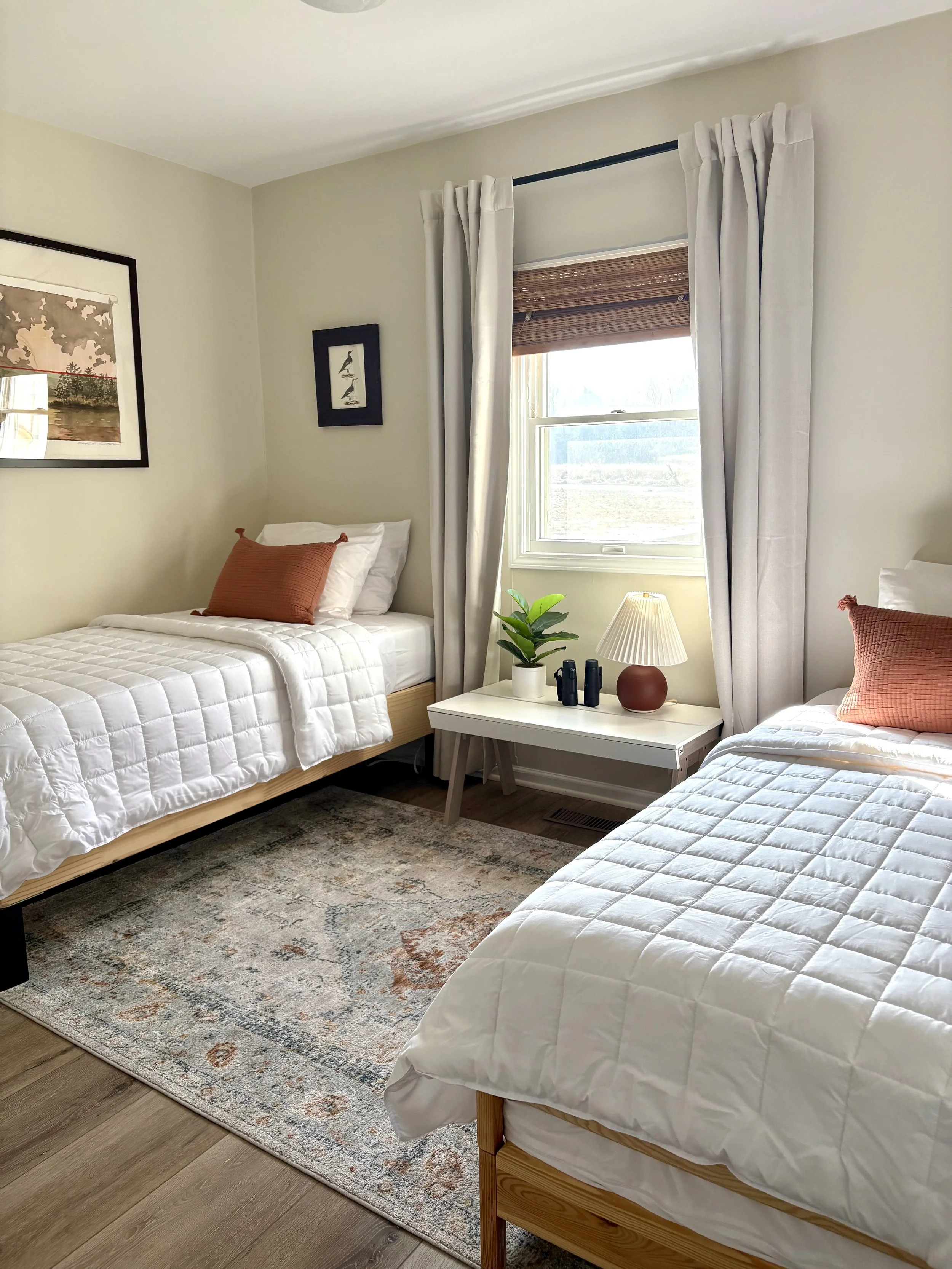 A bedroom with two single beds with white bedding and orange pillows, a window with white curtains, a side table with a lamp, plants, and decorative items, and framed artwork on the walls.