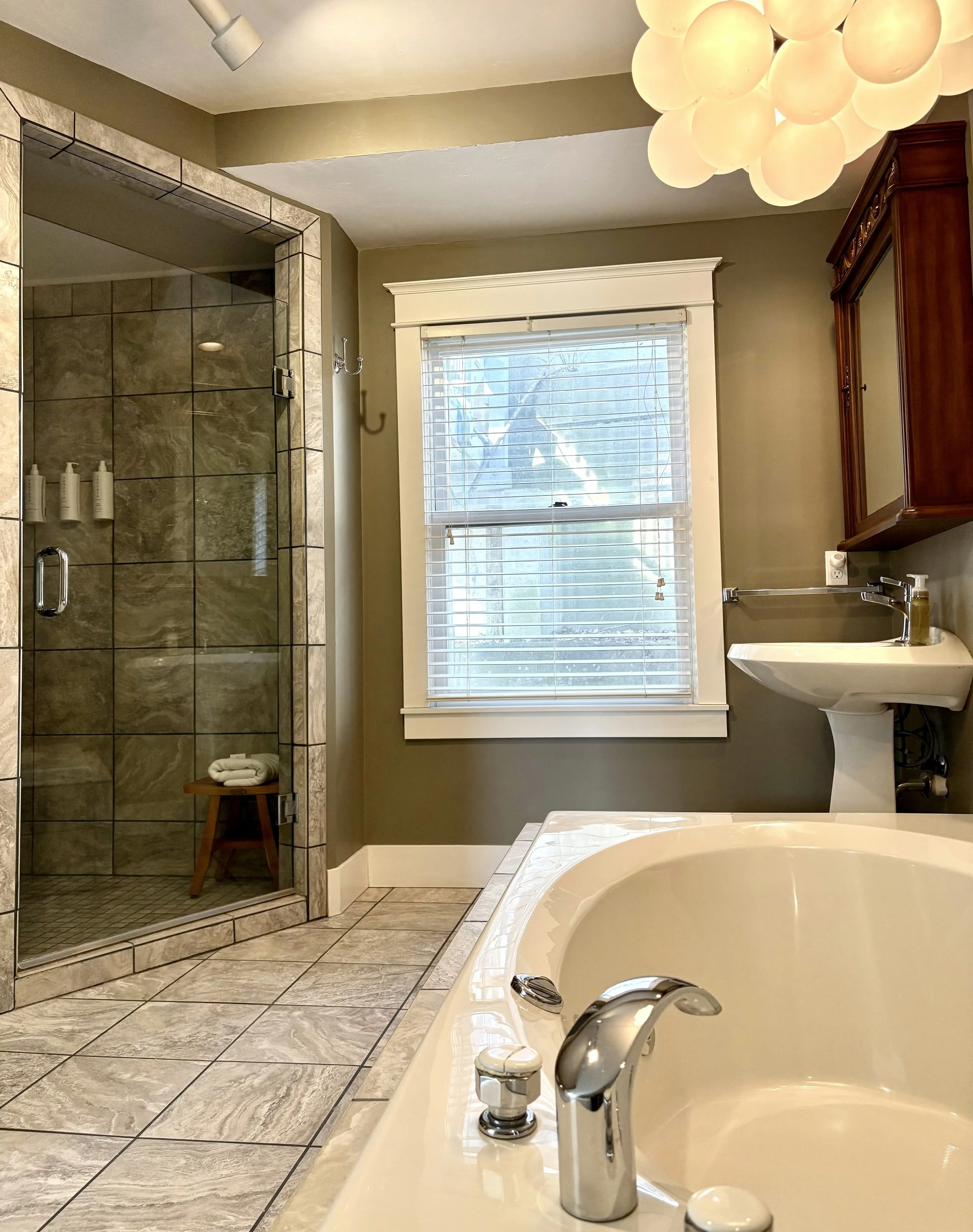 Bathroom featuring a corner shower with glass door, a bathtub, a window with blinds, a pedestal sink, a wooden cabinet, and a unique ceiling light fixture made of round globes.