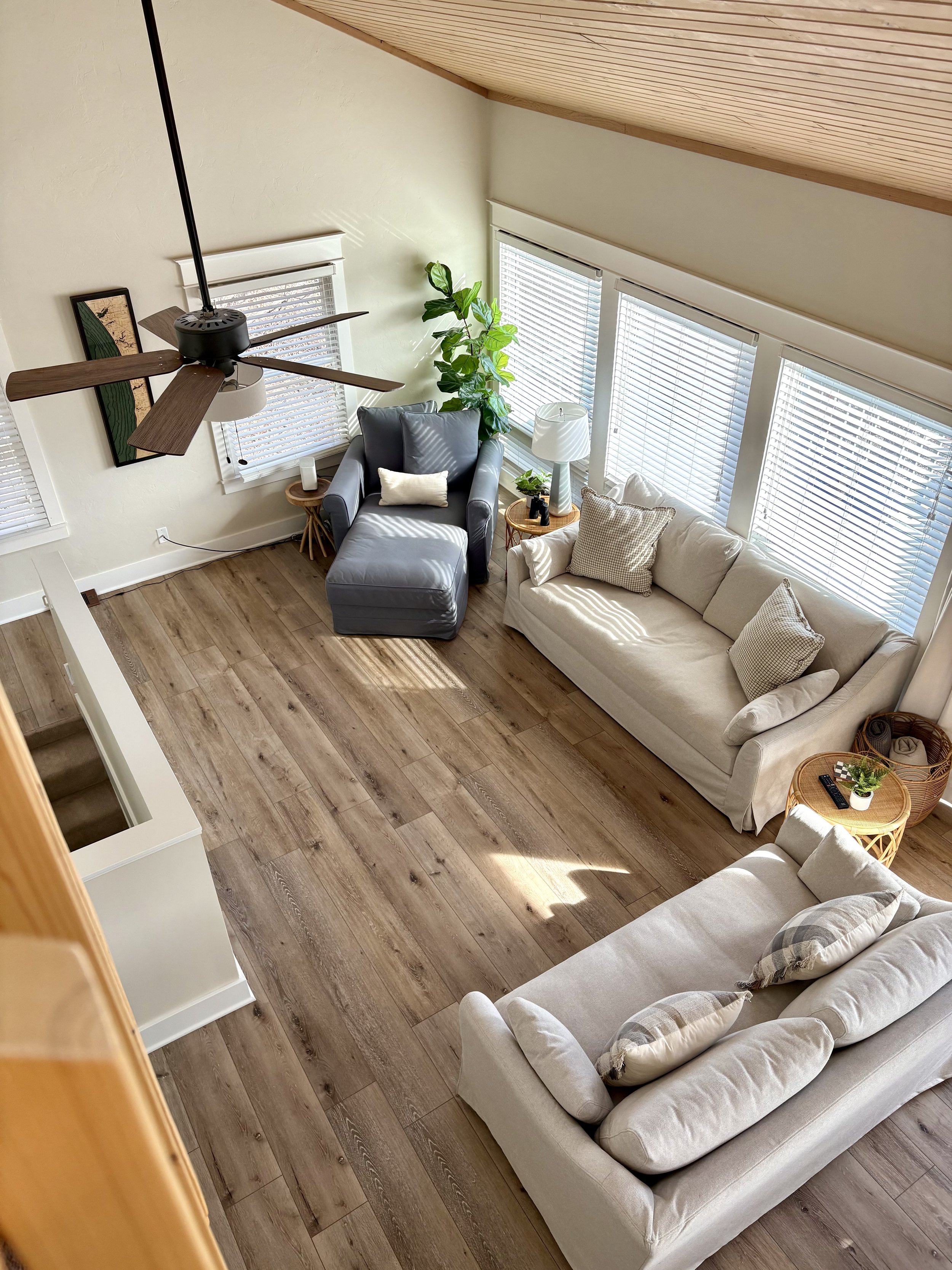 A high angle view of a cozy living room with wooden flooring, featuring a beige sofa with multiple pillows, a gray armchair with an ottoman, and small side tables holding potted plants and remote controls. Large windows with blinds let in natural lig