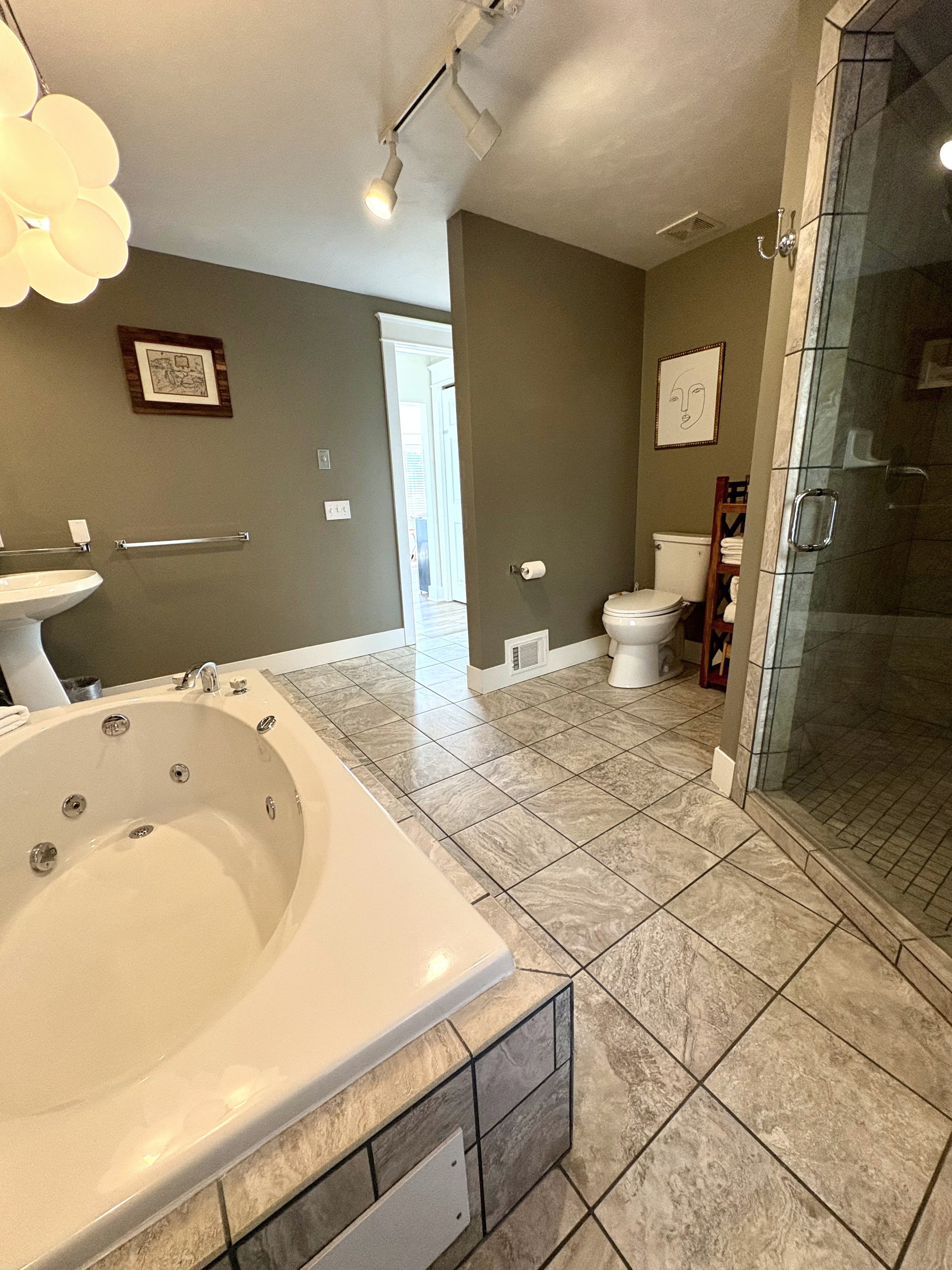 A spacious bathroom with a whirlpool tub, a toilet, and a glass-enclosed shower. The floor and part of the walls are tiled, and the walls are painted a muted green. There is a framed artwork on the wall and a small storage shelf with towels near the 