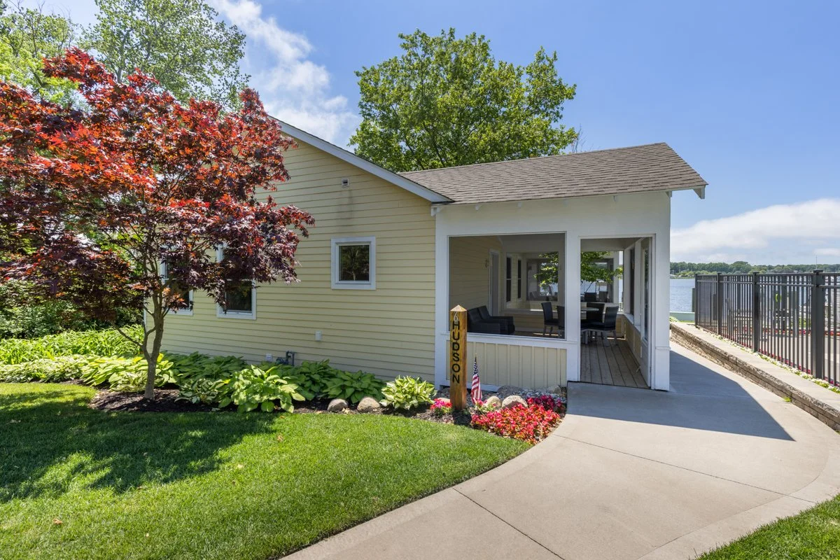 A yellow house with a front porch overlooking a body of water, surrounded by lush greenery, a red-leaved tree, and a landscaped garden with pink flowers.