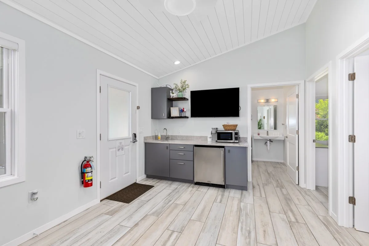 A small kitchenette with a gray cabinet, a microwave, and a mini refrigerator, with a television mounted on the wall overhead and an adjacent bathroom with a sink visible in the background.