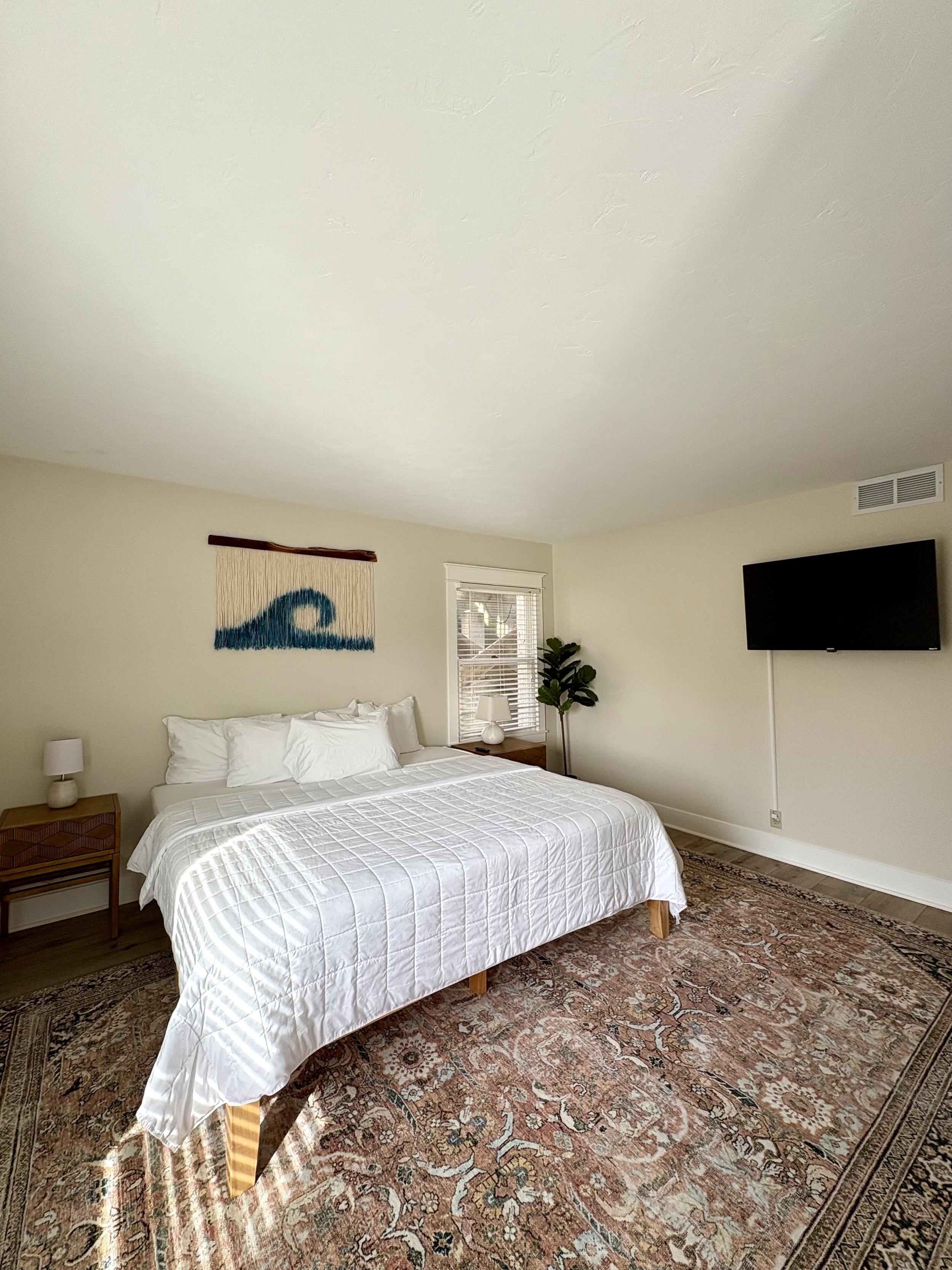 A bedroom with a white bed, a patterned area rug, a wooden nightstand with a lamp, a window with blinds, a potted plant, and a wall-mounted flat-screen TV.