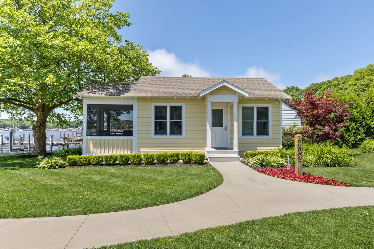 A small, yellow house with white trim, located near a body of water. It is surrounded by green grass, trees, and flowers, with a boat dock visible in the background. A sidewalk leads up to the front entrance and a wooden sign reads 'DURANT'.