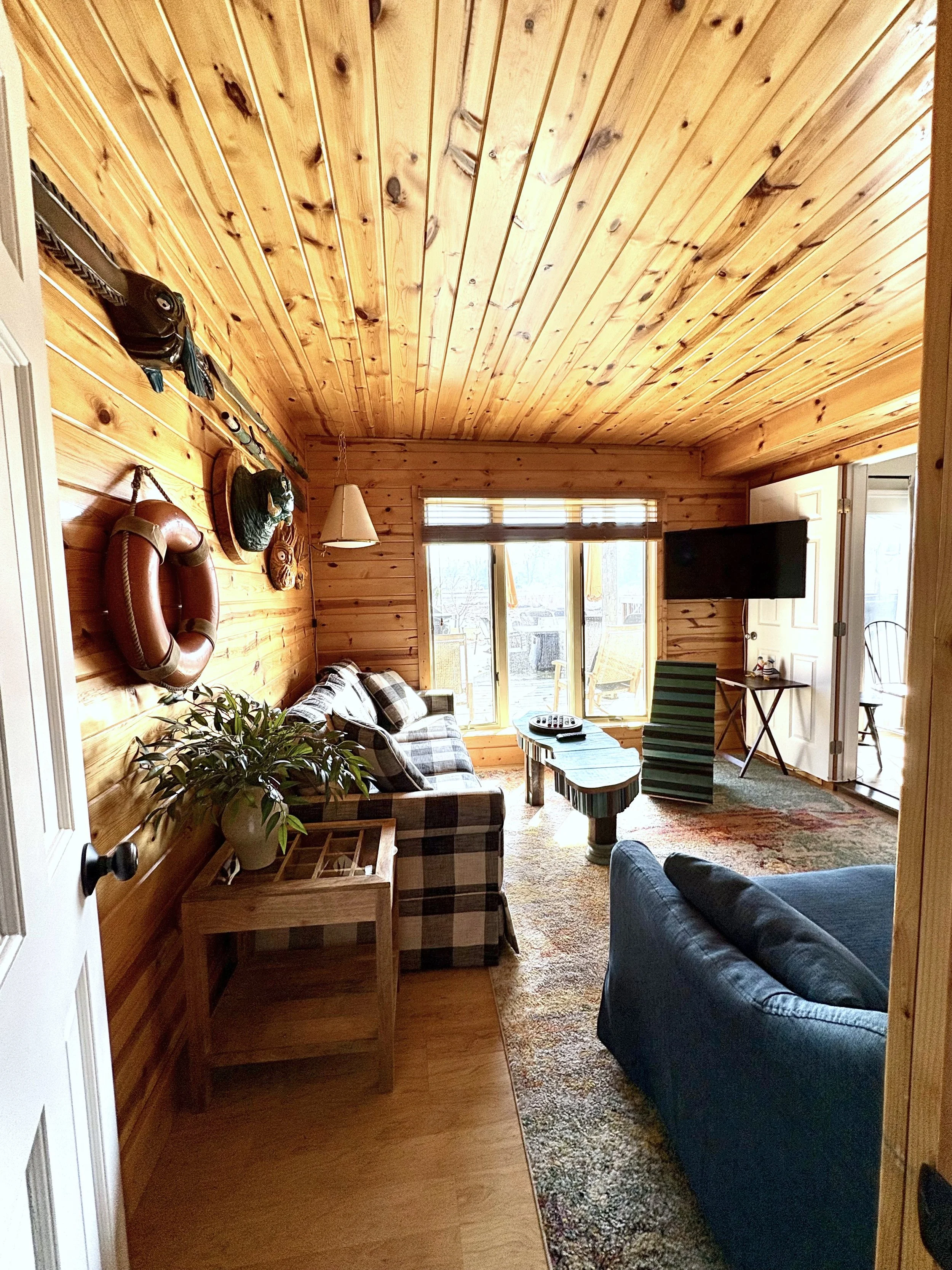 Cozy living room with wood-paneled walls and ceiling, featuring a plaid sofa, a dark blue armchair, a wooden side table with a potted plant, wall-mounted nautical decorations, a wall-mounted TV, and sliding glass doors leading to an outdoor deck with