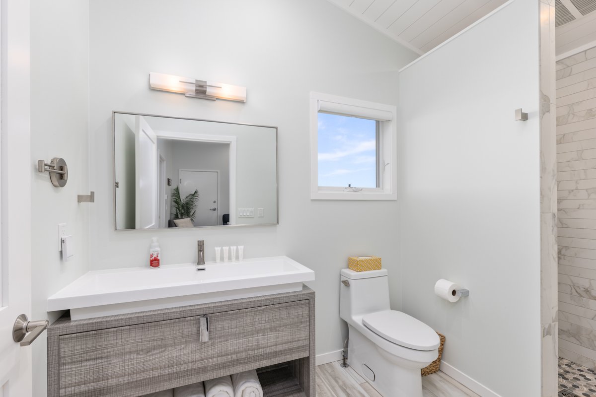 Modern bathroom with a white sink, wooden vanity, large mirror, white toilet with tissue box, window with a view of the sky, small shower area, and minimal decor.