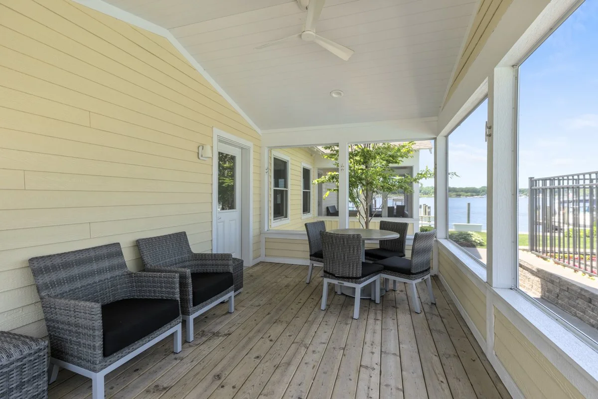 Enclosed porch with wicker chairs, a round table with four chairs, yellow siding walls, large windows showing a lake view, ceiling fan, and wooden deck flooring.