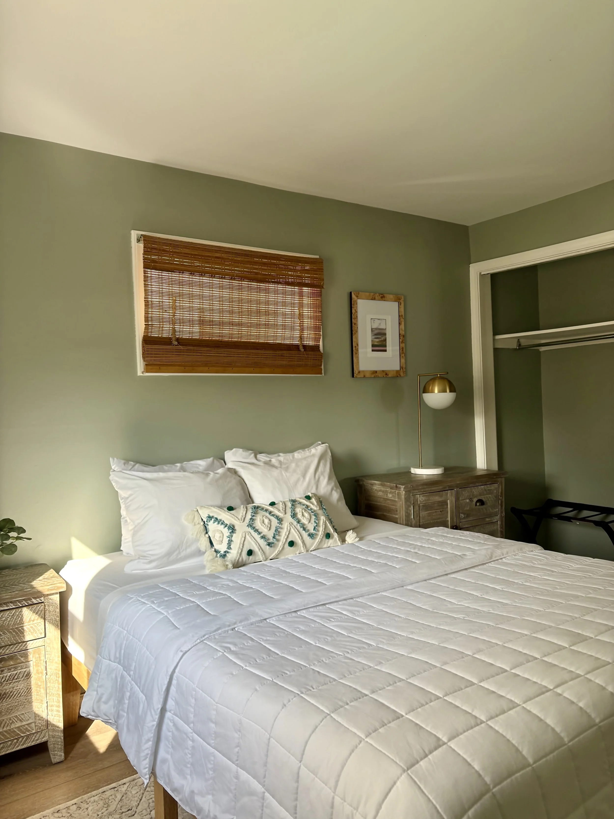 A neatly made bed with white bedding and pillows, a wooden nightstand, a small window with a bamboo blind, framed artwork, and a modern desk lamp in a cozy bedroom with green walls.