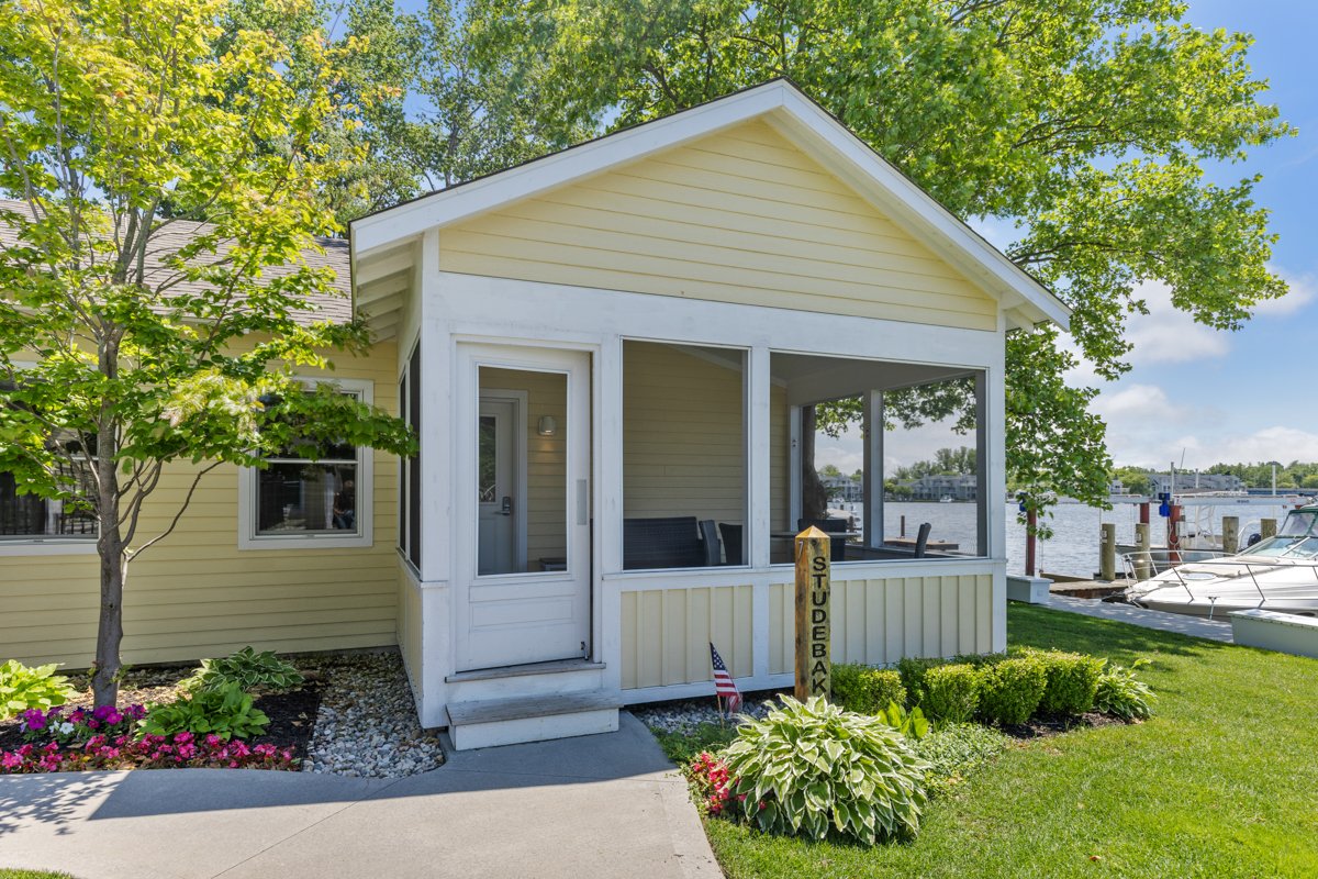 Yellow house with screened porch near a body of water, with boats docked nearby, surrounded by green trees and landscaping.