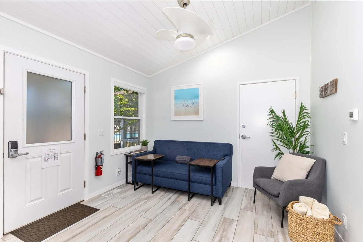 Bright, airy waiting room with gray and blue seating, large window with outdoor view, potted plant, wall art, ceiling fan, and a laundry basket with rolled towels.