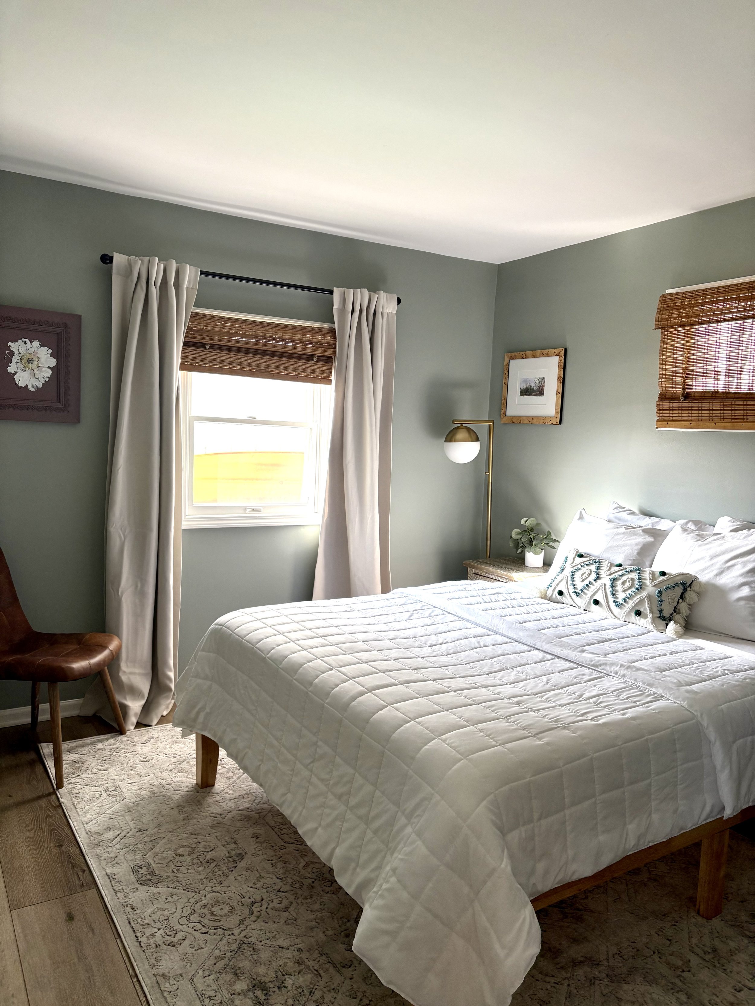 A tidy bedroom with light green walls, a bed with white quilt and pillows, wooden accents, a window with white curtains and bamboo shades, a small side table with a plant, a floor lamp, and framed wall art.