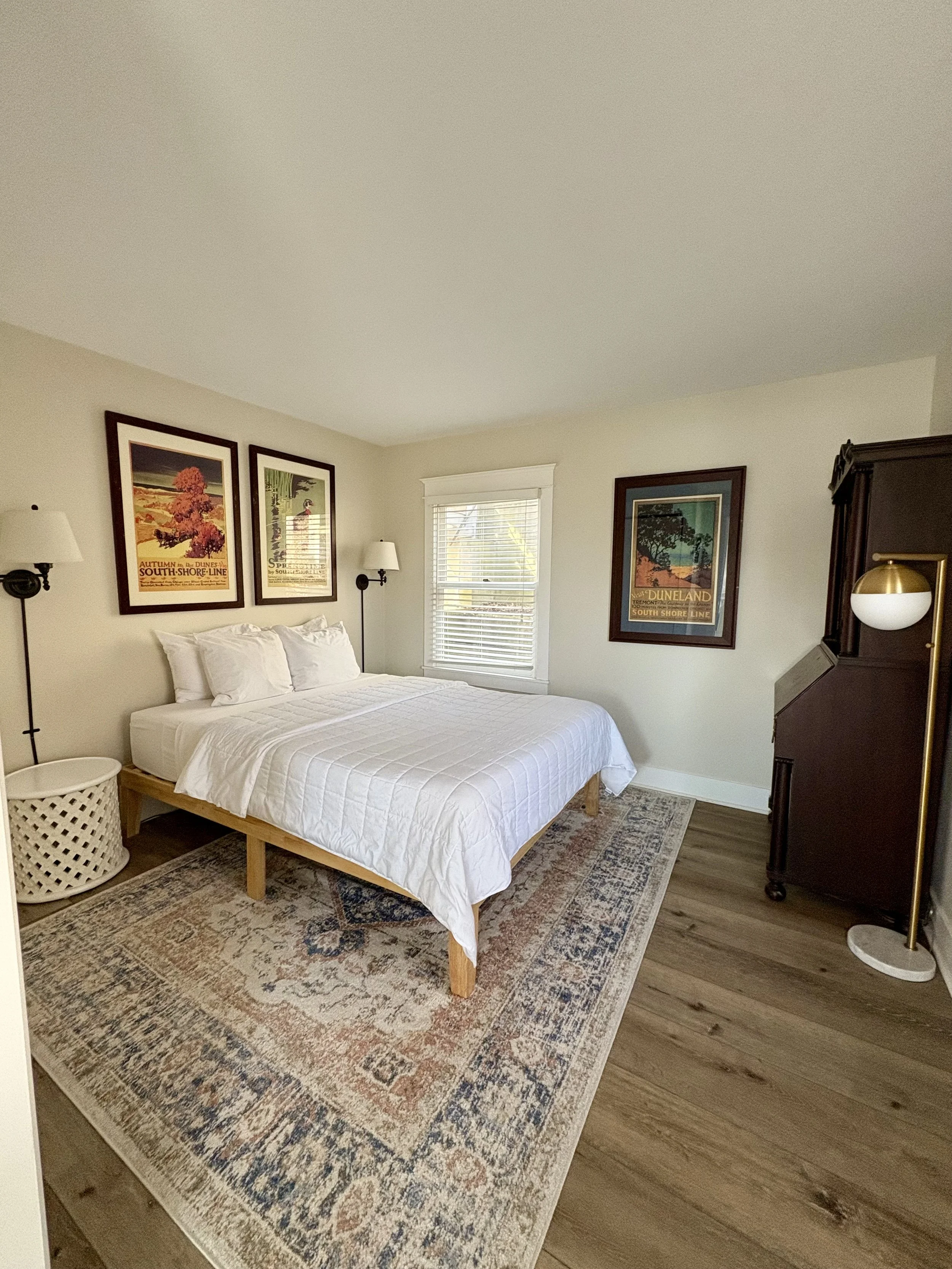 A clean bedroom with a bed covered in white bedding, two pillows, and a wooden bed frame. There are three framed vintage travel posters on the cream-colored walls, two beside the bed and one to the right. A window with white blinds is centered on the