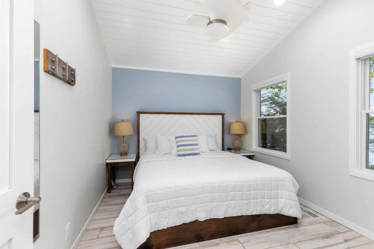 A bedroom with a white bed, a wooden headboard, two nightstands with lamps, and two windows showing trees outside.