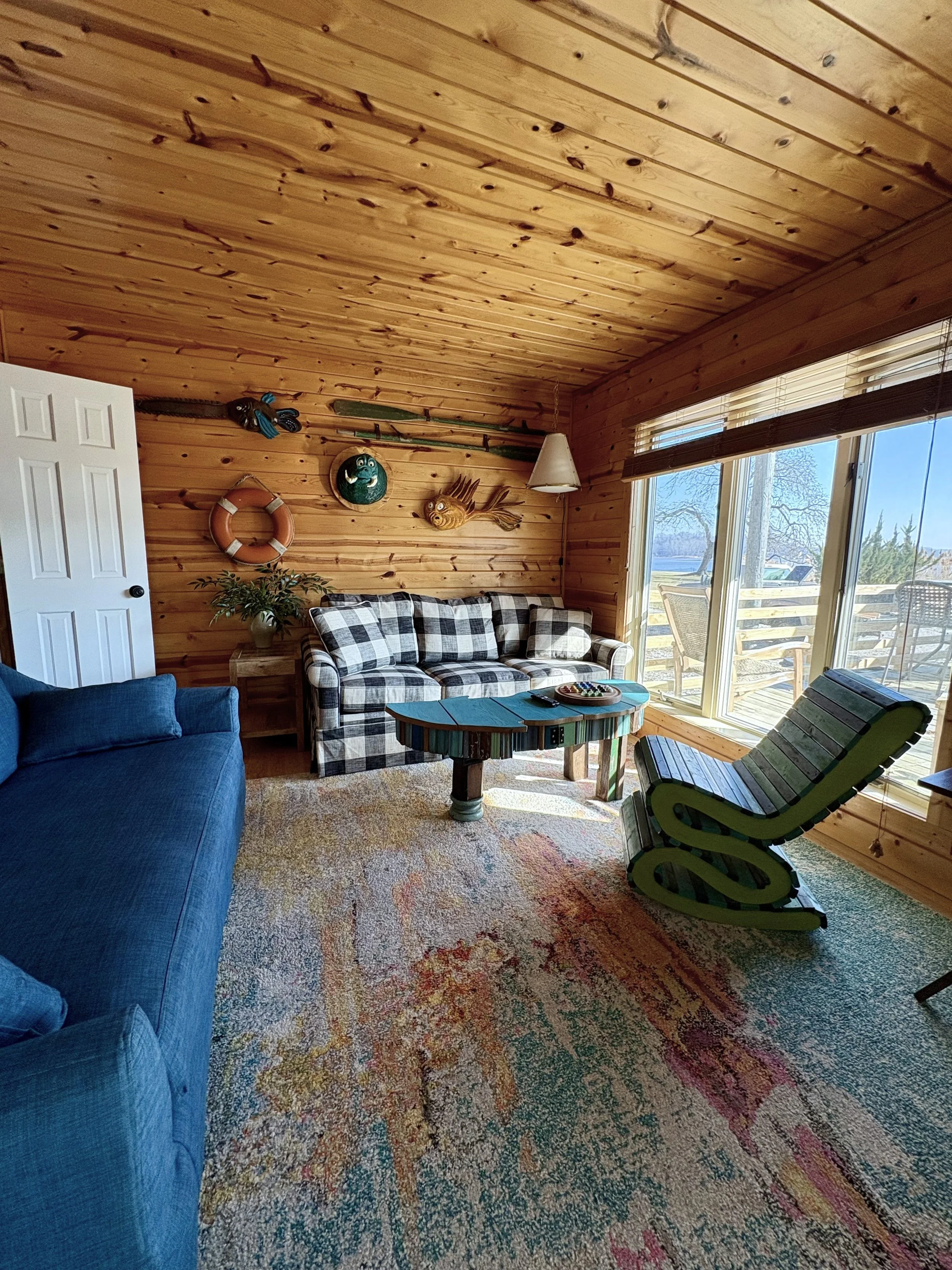 Sunlit living room with wood-paneled walls and ceiling, featuring a checkered sofa, a colorful patterned rug, a blue armchair, a green recliner, and nautical decorations including fishing poles, a lifebuoy, a fish wall art, and a potted plant near a 