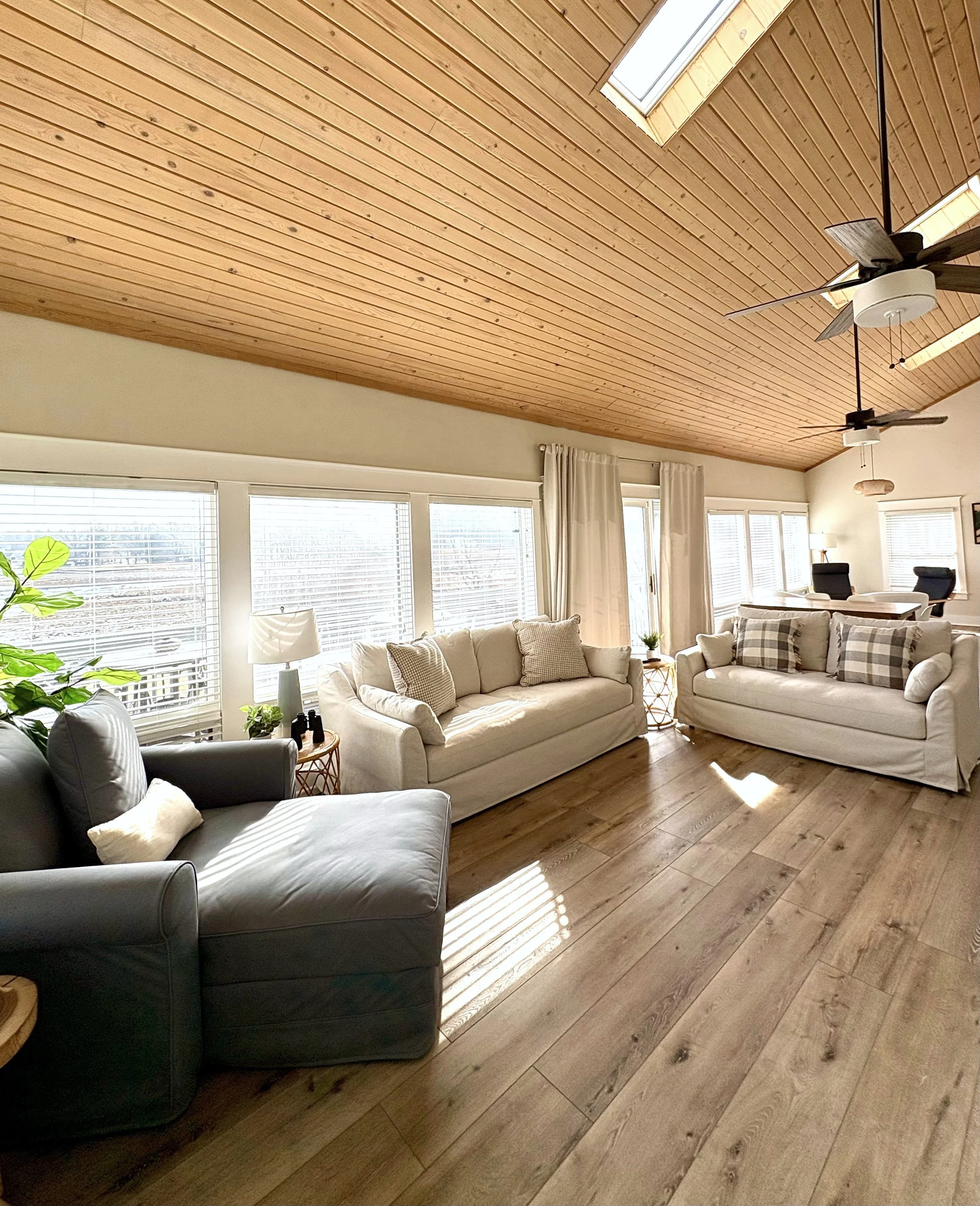 A bright living room with a wooden vaulted ceiling, large windows with white blinds, and three sofas with neutral-colored cushions. There are potted plants, side tables, and ceiling fans.