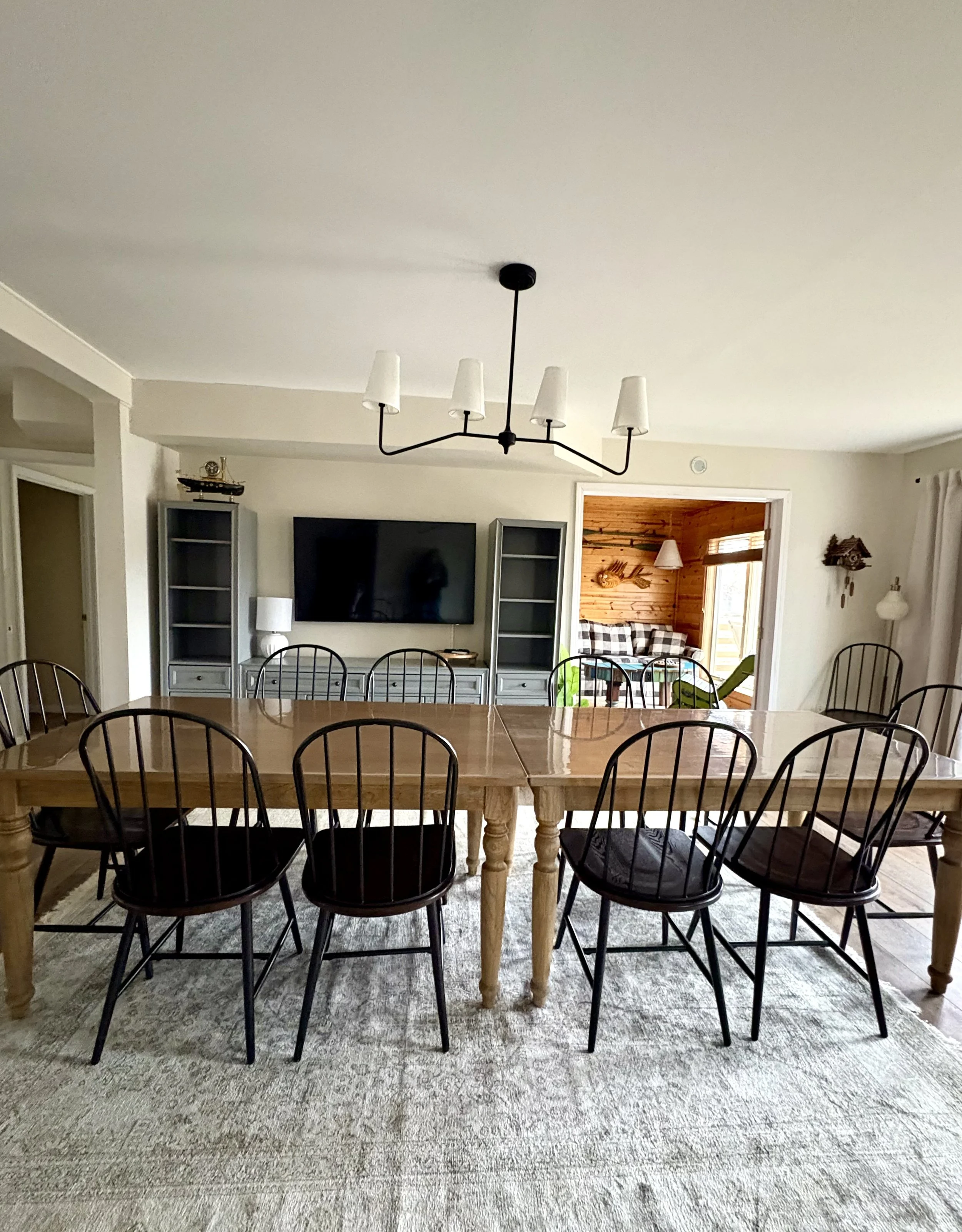 Dining room with a large wooden table and eight black chairs, a wall-mounted television, and a sitting area with a bookshelf and lamp visible through an open doorway.