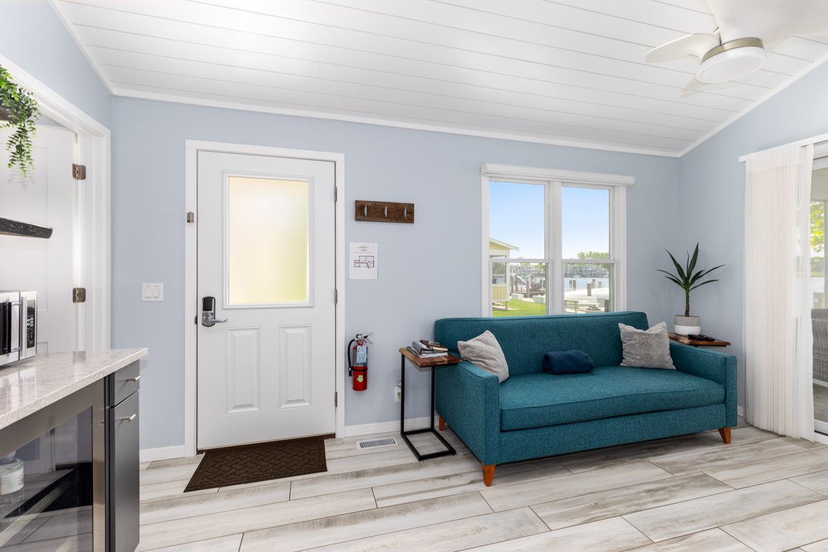 Living room with a teal couch, side tables, a potted plant, large windows, and a door, with light blue walls and wood-look flooring.