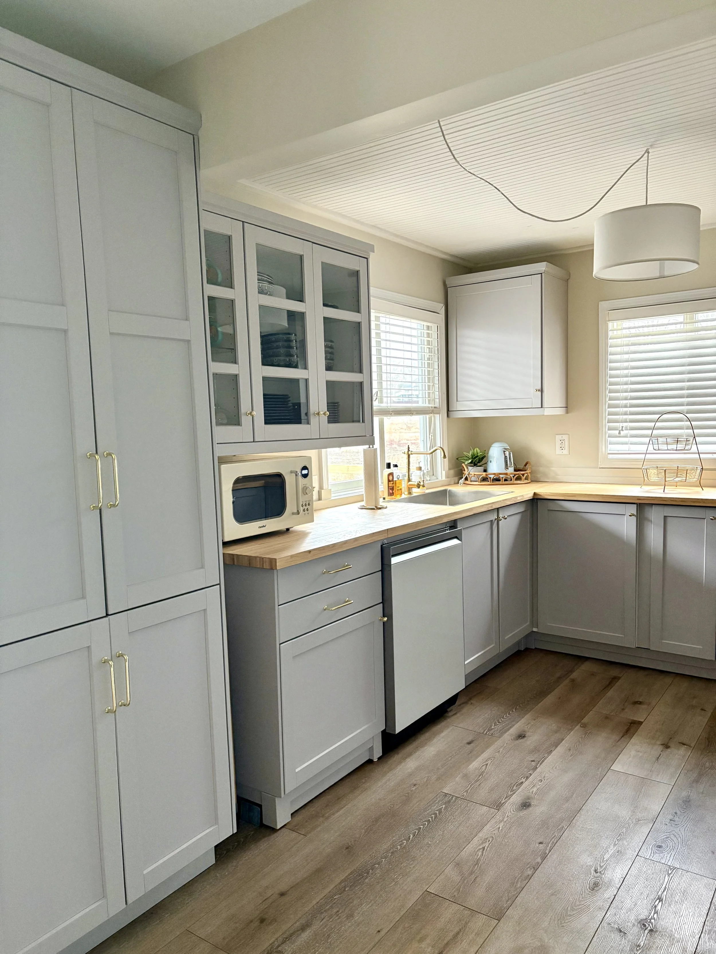 Kitchen with light gray cabinets, wooden countertops, and hardwood flooring. There are two windows above the sink, a microwave on the counter, and a hanging light fixture.