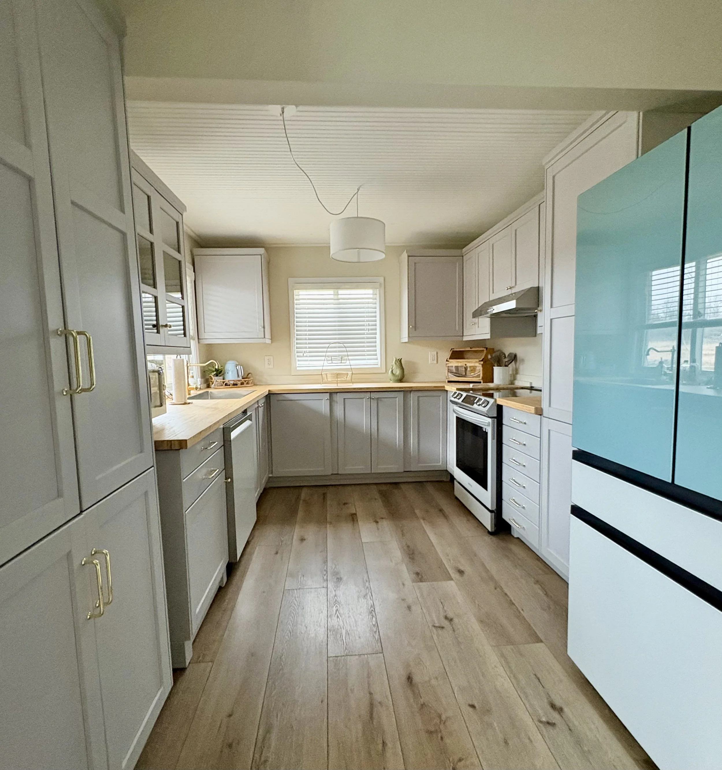 A modern kitchen with gray cabinetry, light wood countertops, and a light wood floor. There's a window above the sink, and a turquoise refrigerator on the right side. The kitchen has a white ceiling with a light fixture hanging from it.