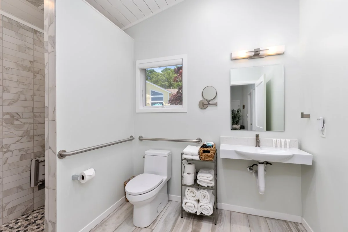 Bathroom with a toilet, a small window, a wall-mounted mirror, a sink with toiletries, a metal shelving unit with towels and toilet paper, and a shower area with marble tiles.