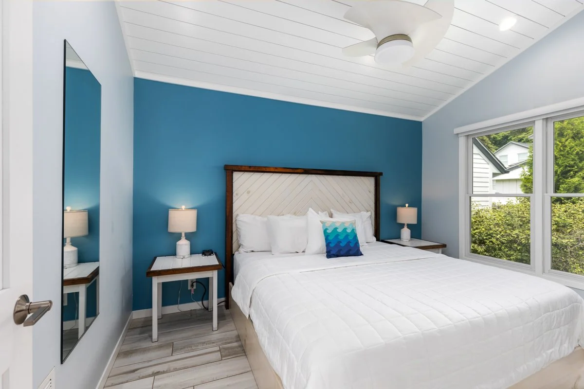 A bedroom with a white bed, blue accent wall, two bedside tables with lamps, a large window with a view of greenery, and a ceiling fan.