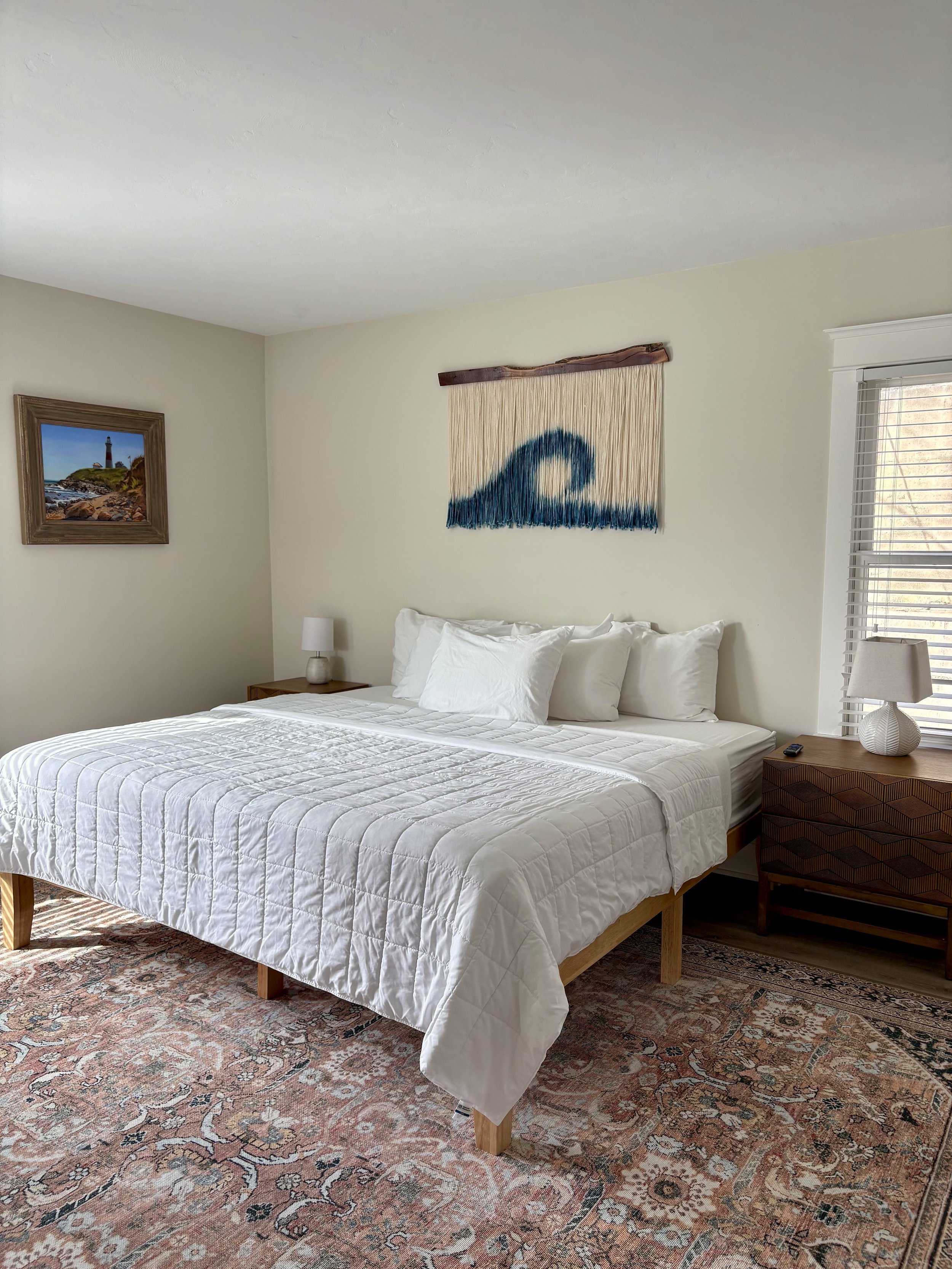 A neatly made bed with white bedding and multiple pillows in a bedroom with a decorative rug, wall art, a window with blinds, and bedside tables with lamps.
