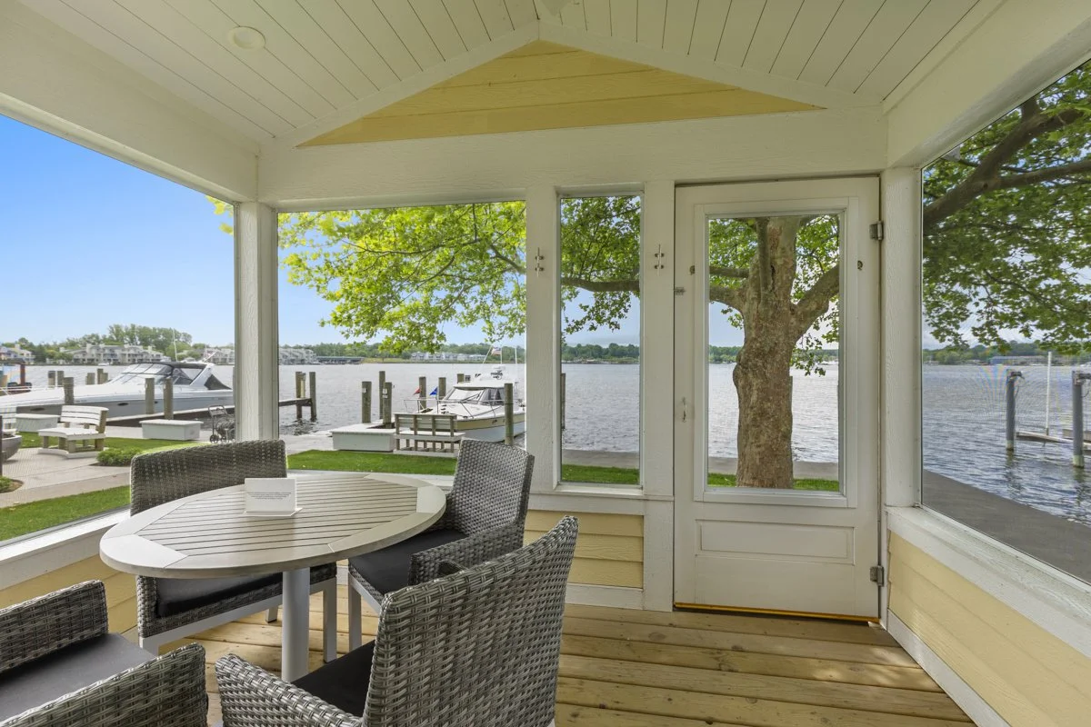 Sunroom with a view of a waterfront, boats docked at the pier, a large tree outside, wicker chairs around a round table, and a door leading outside.