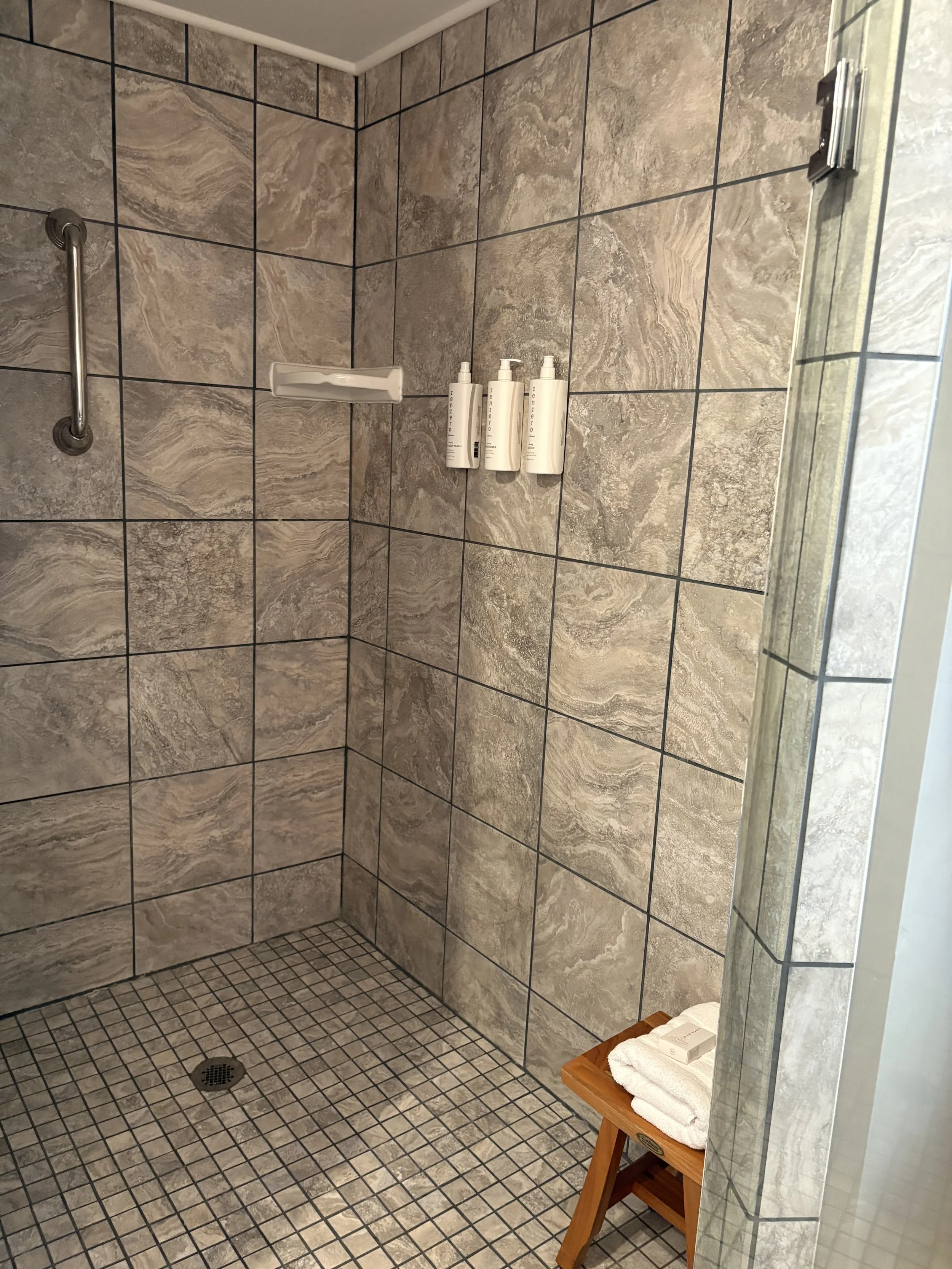 A shower with beige and gray tiled walls and floor, a metal grab bar, three white bottles on the wall, a soap dish, and a small wooden stool with folded towels in the corner.