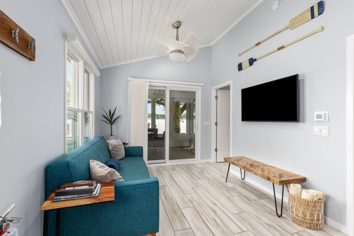 Living room with light blue walls, a teal sofa with pillows, a wooden side table with magazines, a large flat-screen TV on the wall, three paddles mounted above the TV, a sliding glass door leading to a screened porch with a lakeside view, a potted p