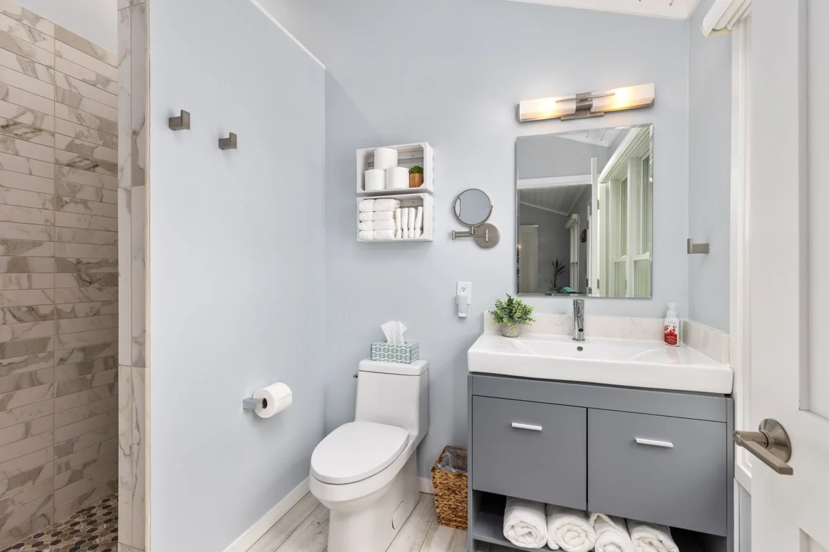 Modern bathroom with light blue walls, a toilet, gray vanity with a white sink, a large mirror, wall-mounted light fixture, and glass shower with marble tiles.