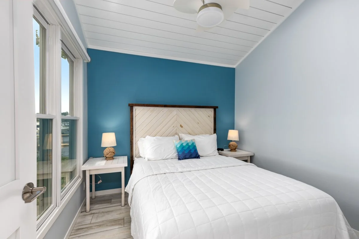 A bedroom with white walls and a blue accent wall behind the bed, featuring a white quilted bedspread, pillows, and a decorative blue pillow. Two white nightstands with lamps are on either side of the bed. The room has three windows with white trim a