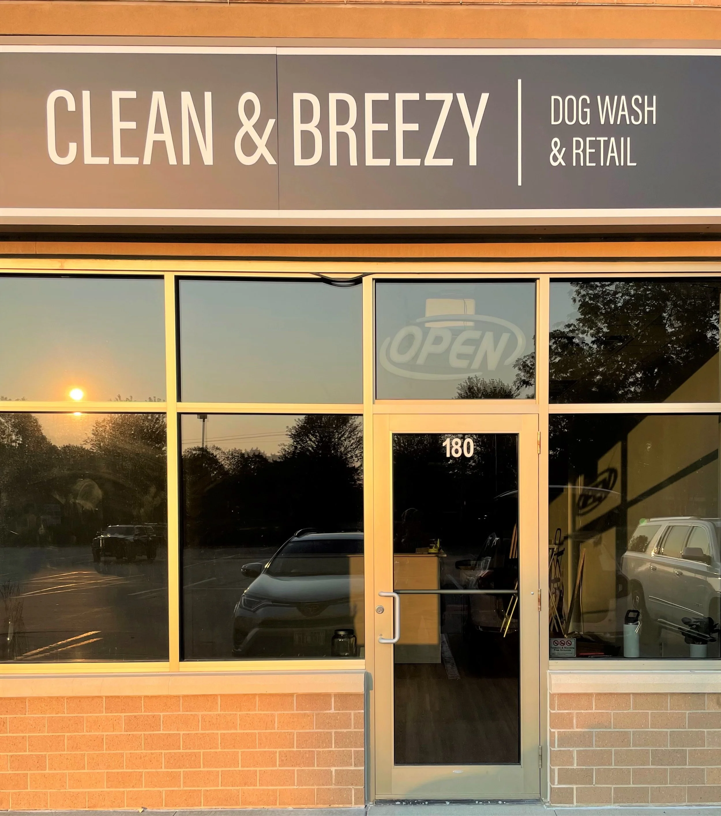 Sign for 'Clean & Breezy' dog wash and retail store, with glass door and window reflecting the sunset, trees, and parked cars outside.