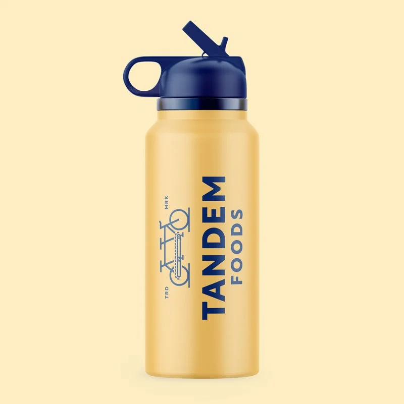 Gold water bottle with blue vertically stacked Tandem logo. Bottle with blue cap on tan background.