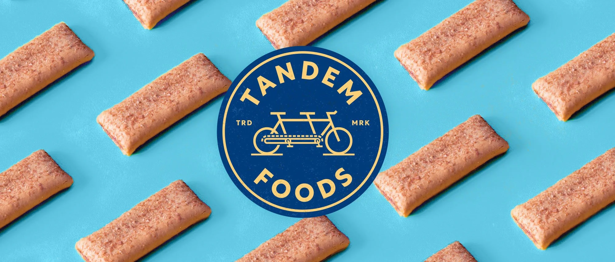 Blue and gold Tandem Foods emblem with tandem bike artwork and pattern of nutrition bars on blue in background.