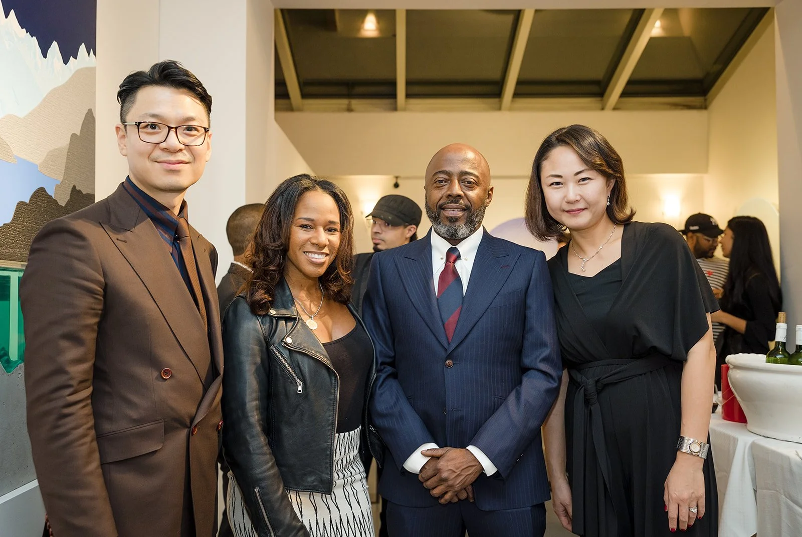 Group of four diverse adults, two men and two women, dressed in formal attire, standing together in an indoor event space with other people in the background.