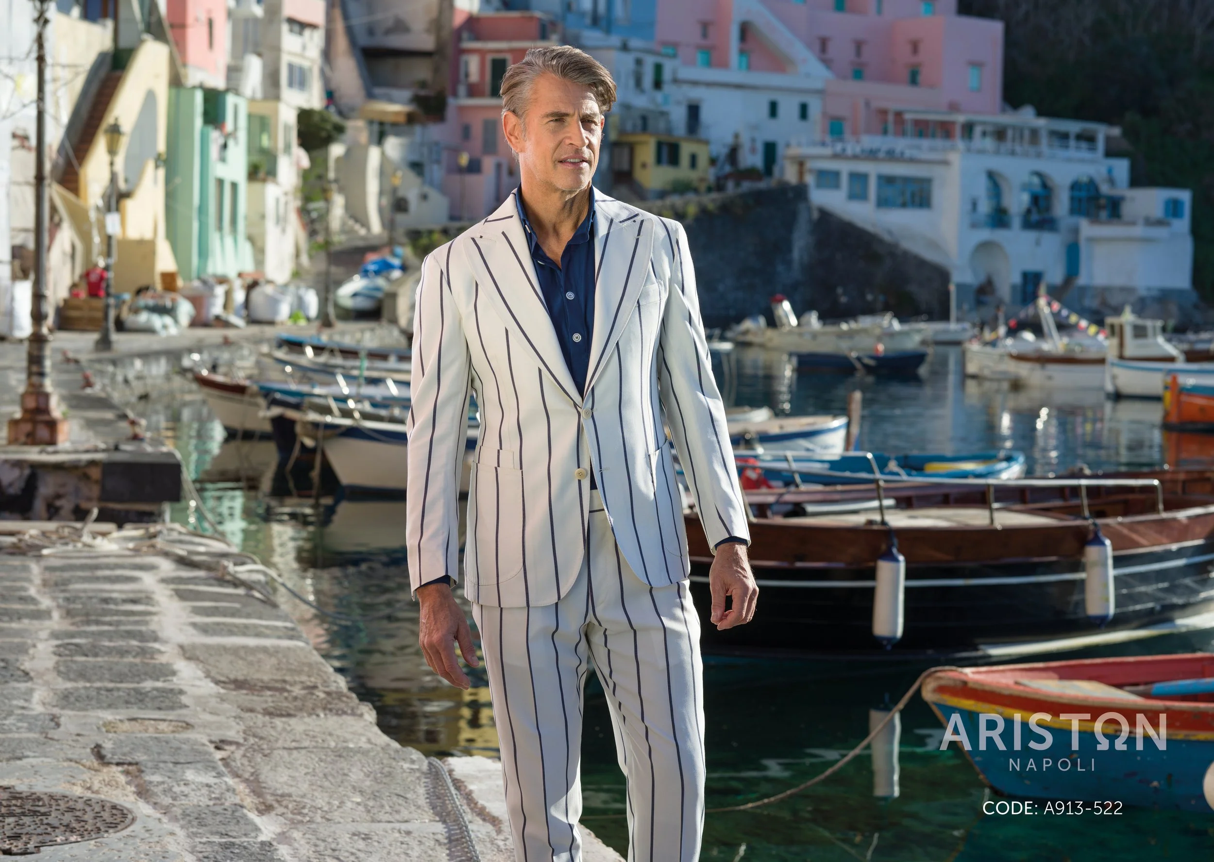 A man in a white suit with navy pinstripes walking along a harbor with colorful buildings and boats in the background.