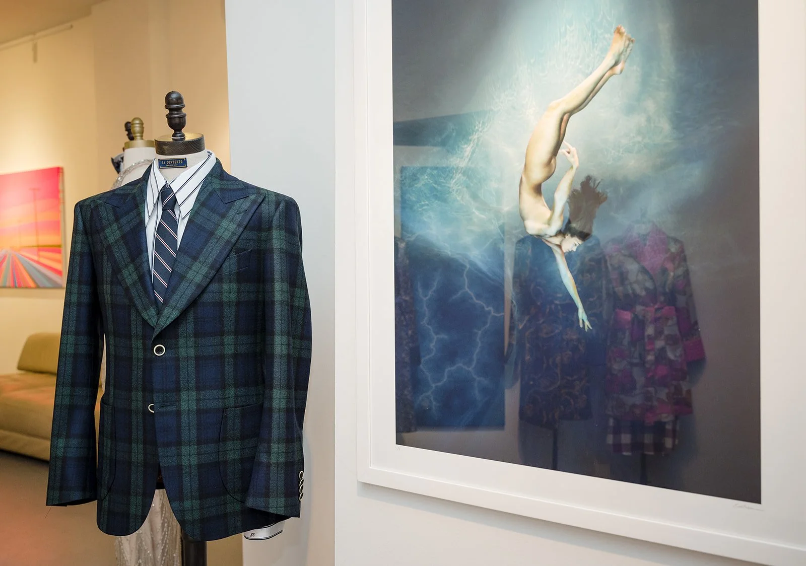 A mannequin dressed in a plaid suit with a white shirt and striped tie, positioned near a large framed artwork of a woman underwater. The art piece features a woman with dark hair submerged in water, surrounded by lightning and stormy elements.