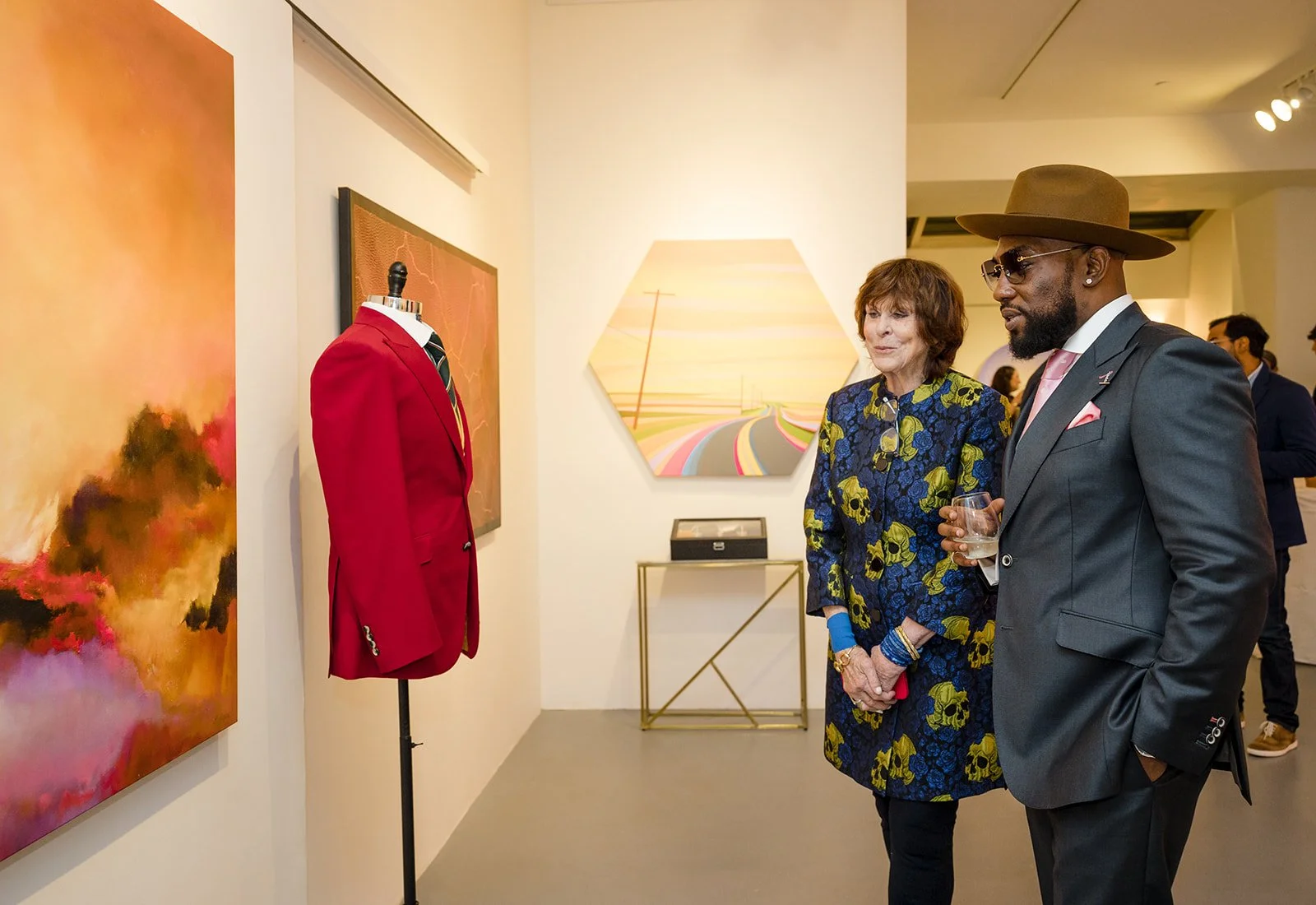 Two people viewing art in a gallery, with one wearing a floral dress and the other in a dark suit with a brown hat, holding a glass of water. Artworks and a mannequin with a red blazer are visible.
