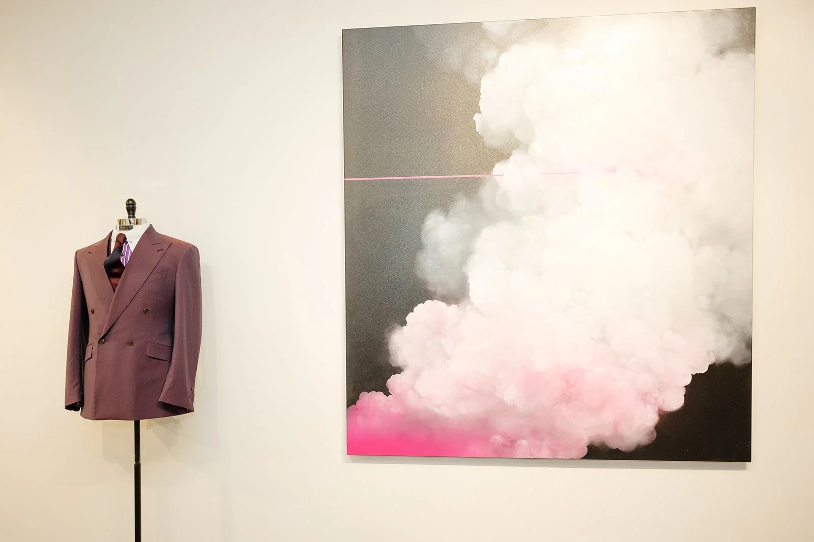 A burgundy men's suit on a mannequin next to a modern abstract painting of clouds with pink, white, and black colors on a white gallery wall.