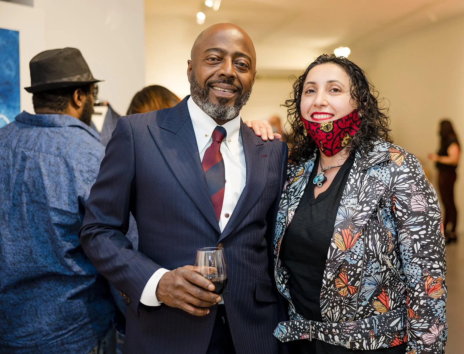Man in a navy pinstripe suit holding a glass of red wine smiling next to woman in a floral butterfly patterned jacket and a red face mask, at an indoor social event.