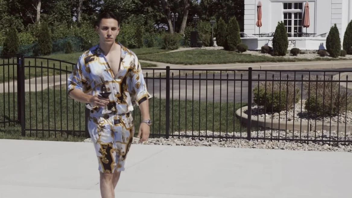 A young man in a patterned shirt and shorts walking outside on a sidewalk, holding a glass of red wine, with a residential house and garden in the background.